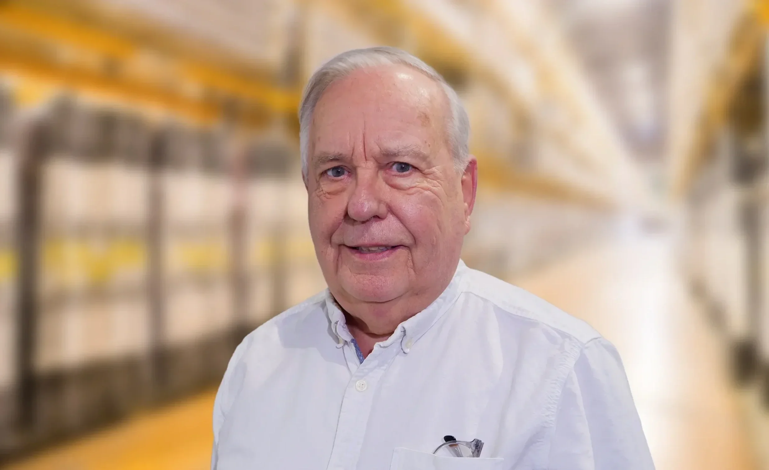 A headshot of a CM Loney Company Secretary called Mervyn Hall. An older man with grey hair, wearing a white shirt with glasses in pocket, standing in a warehouse with shelves filled with boxes in the background.