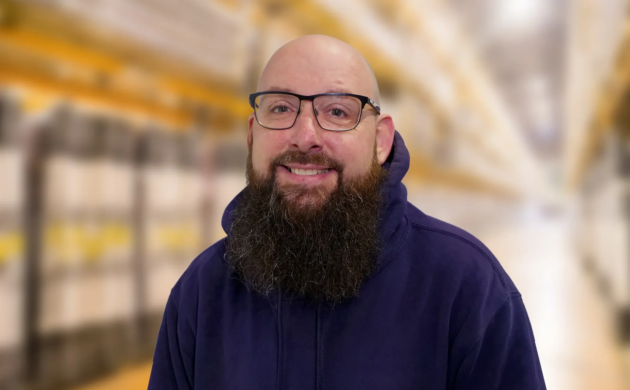 A headshot of a CM Loney electrician called Andy Whiting. A man with glasses and a full beard smiling, standing in a warehouse with blurred shelves in the background.