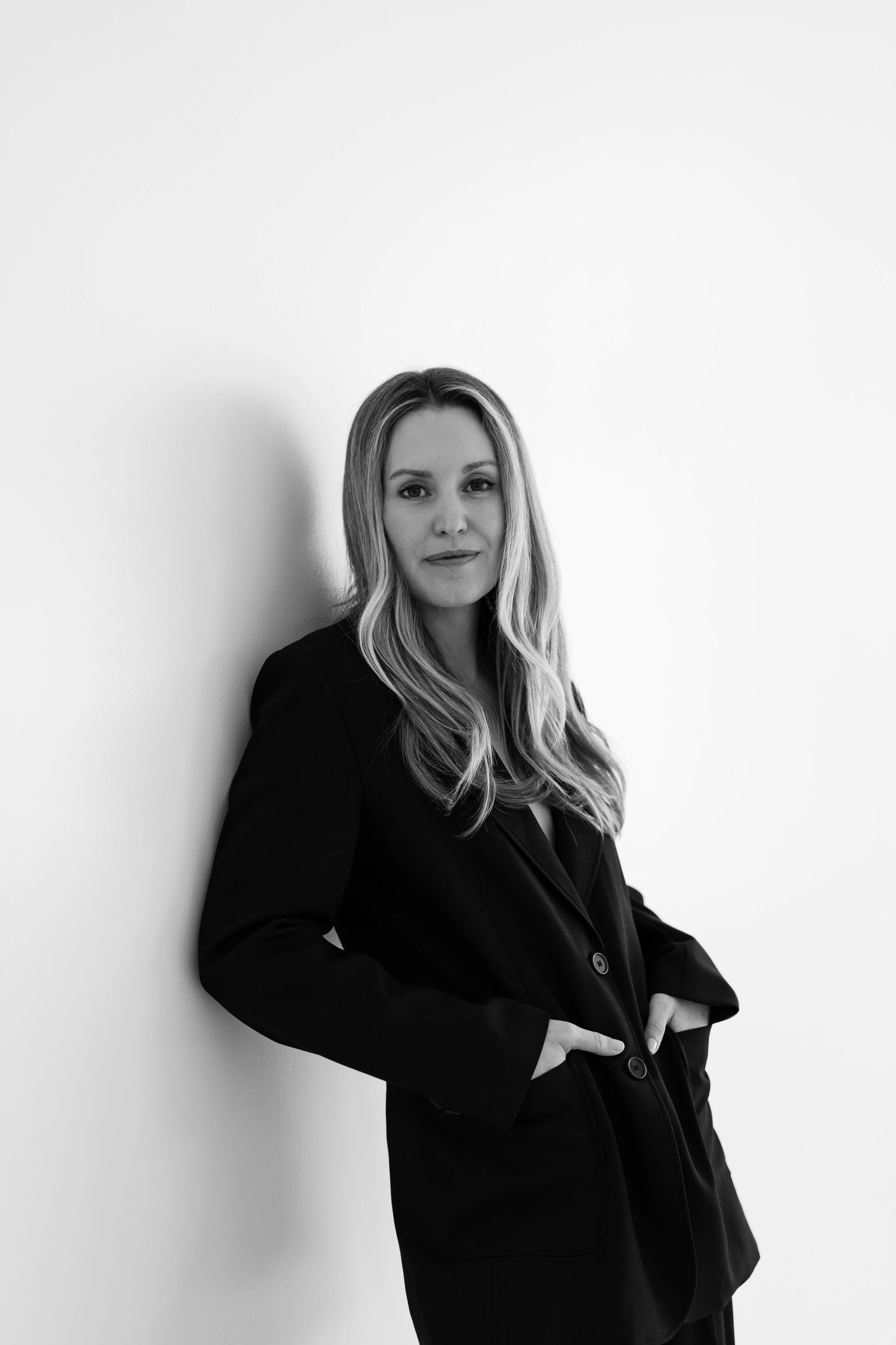 A woman with long wavy hair wearing a black blazer, standing against a plain white wall.