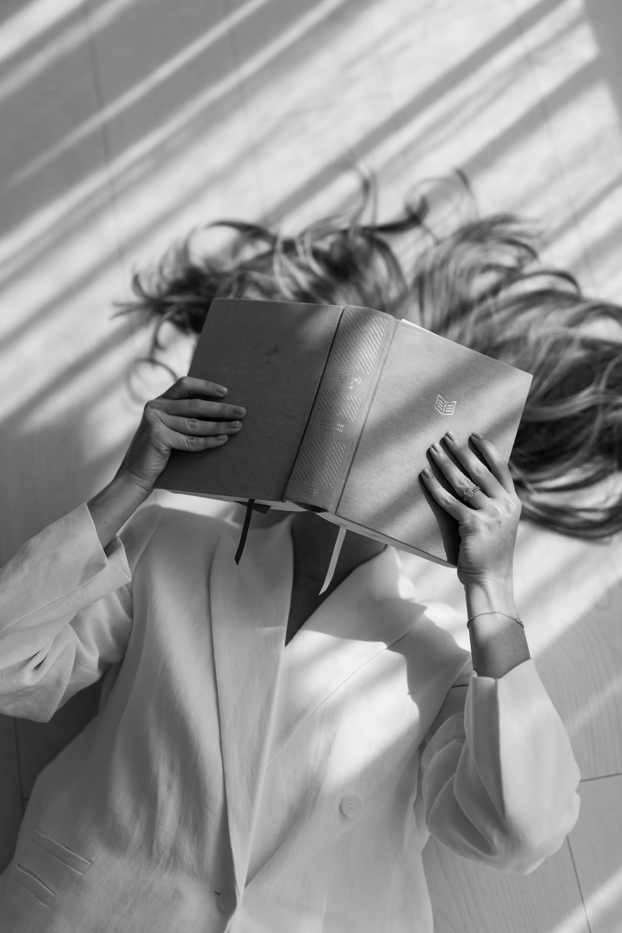 A woman in a white blazer lying on the floor, holding a large book over her face, with sunlight creating striped shadows.