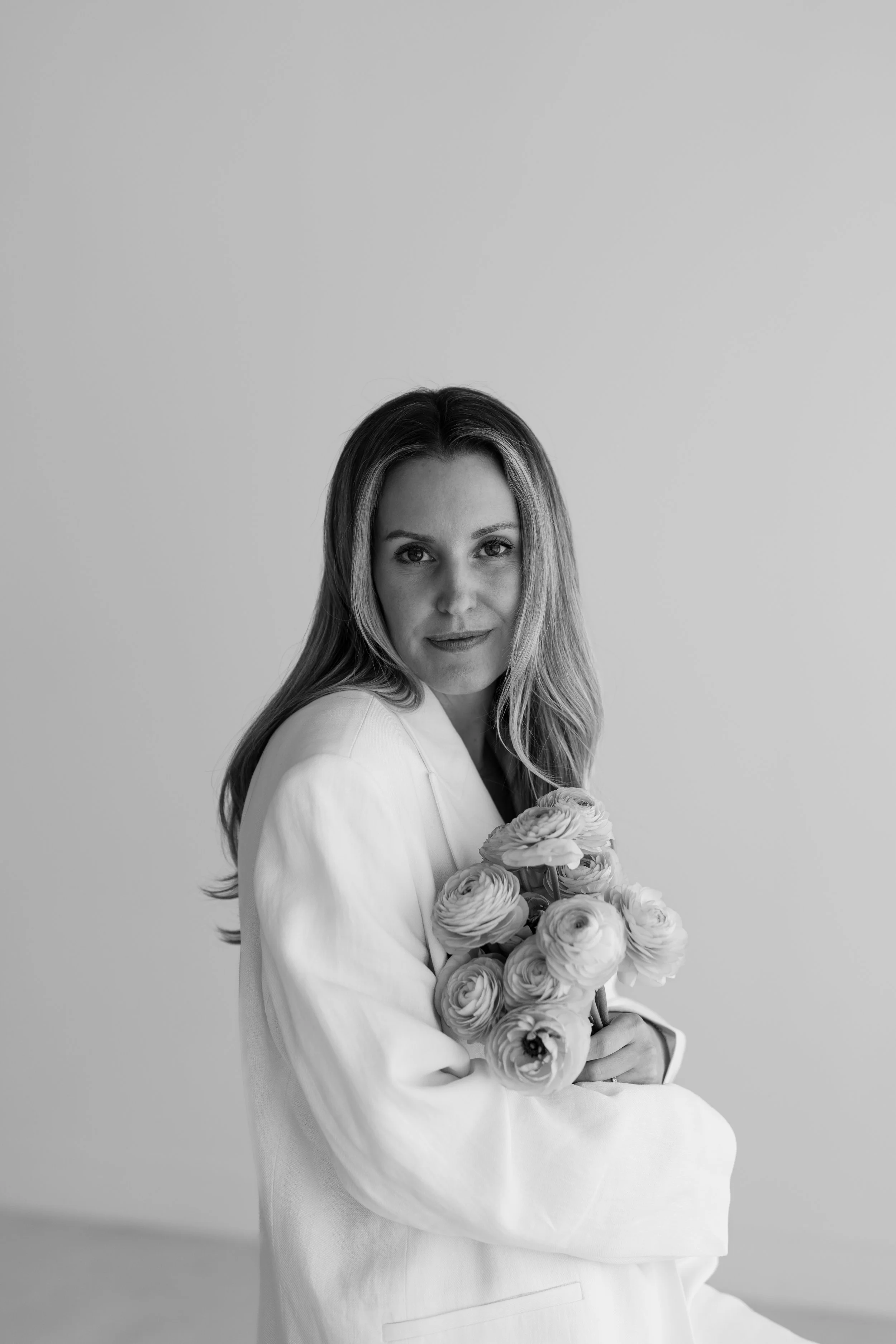 A black and white photo of a woman with long hair holding a bouquet of flowers, wearing a white top or jacket, looking at the camera with a neutral expression.
