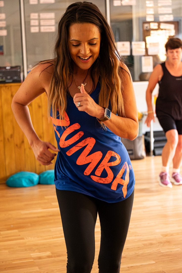 A woman with long brown hair smiling and wearing a blue and orange workout tank top that says "YOGA" in orange, black leggings, and a smartwatch, participating in a fitness class indoors with wooden floors and a woman in black workout clothes in the background.