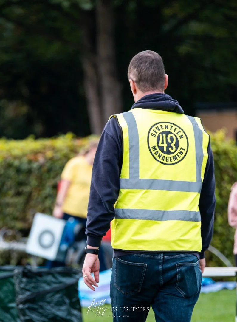 A man wearing a yellow safety vest with the words "Seventeen Management" and the number 43 on the back is standing outdoors, facing away from the camera, with trees and blurred people in the background.