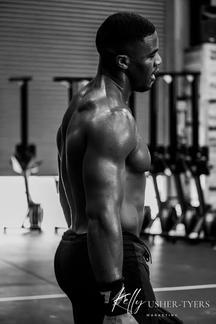 A muscular man with sweat on his body, standing in a gym, shirtless, with his eyes closed and smiling.