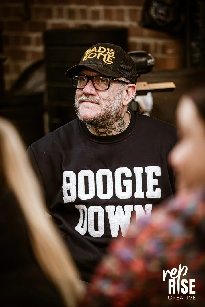 A man with glasses, a gray beard, and tattoos wearing a black cap with 'ADTO THE BOC' written on it, and a black sweatshirt with 'BOOGIE DOWN' printed on it, sitting indoors with a brick wall background.
