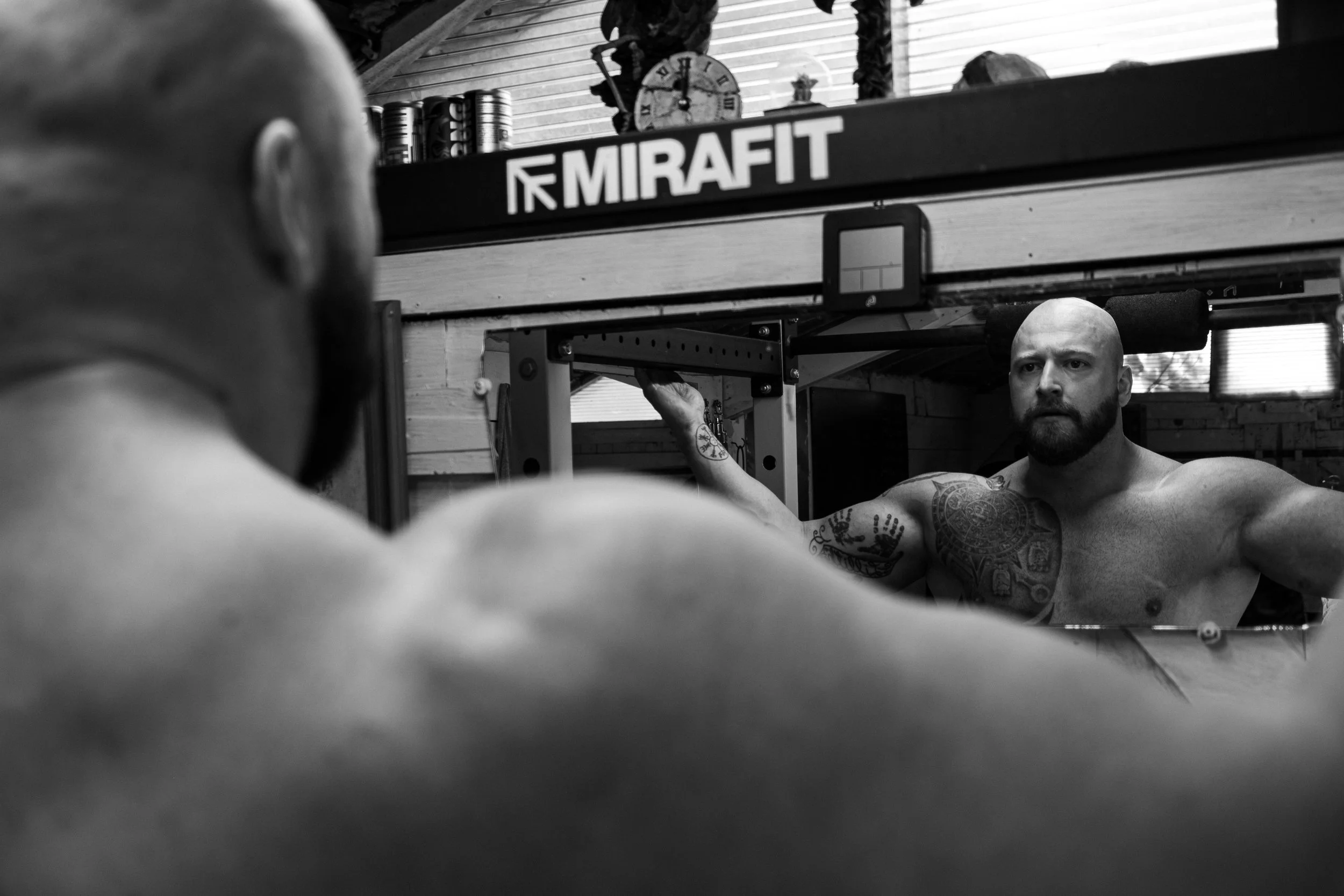 A shirtless man with tattoos looking at himself in a mirror, holding a barbell, with a gym background and the word 'MIRAFFIT' on the wall in black and white.