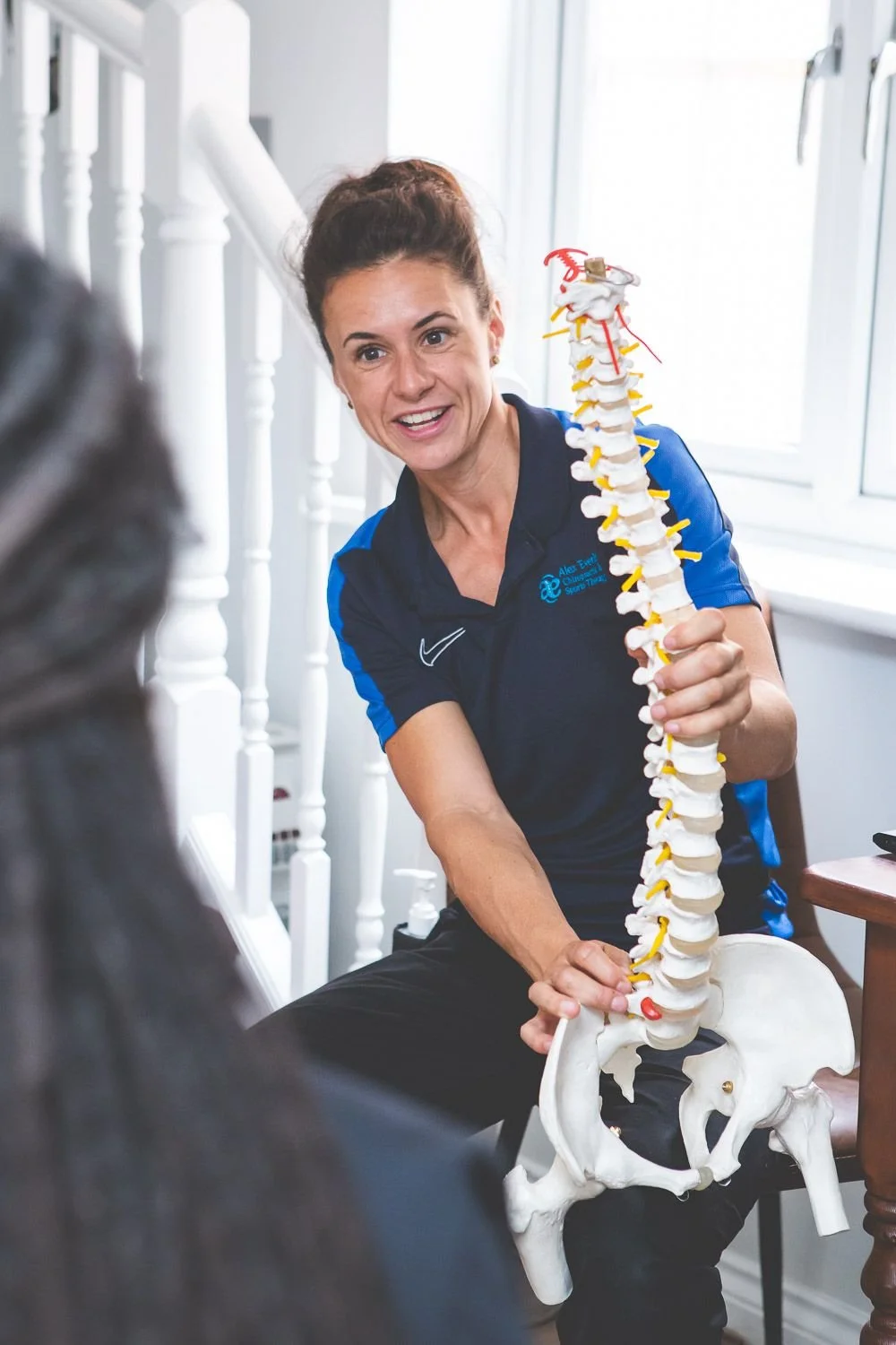 A woman is holding a plastic anatomical model of the human spine and pelvis, demonstrating or explaining it to others.
