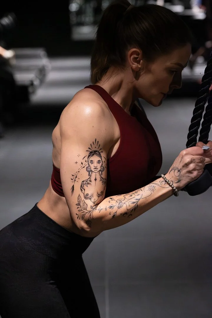 A woman with a ponytail and tattoos on her arm and side, dressed in a maroon sports top and black leggings, working out at a gym, holding a kettlebell.