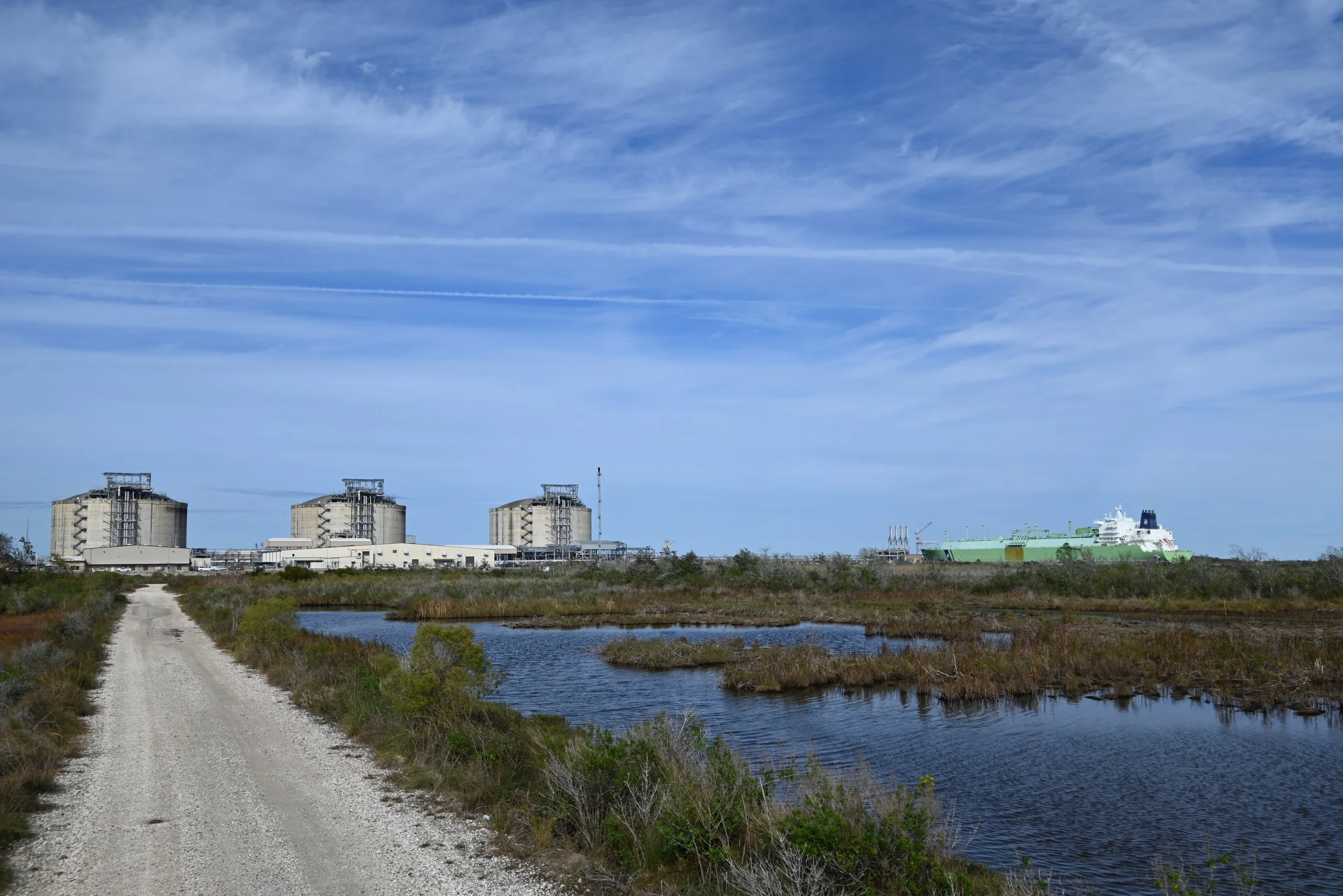 A landscape featuring a gravel path leading towards industrial buildings with large tanks, a waterway on the right, and a ship in the background under a partly cloudy sky.