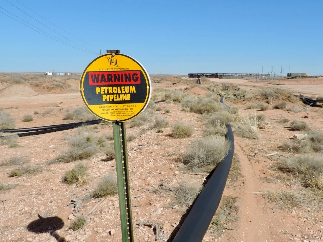Warning sign indicating a petroleum pipeline in a desert area with sparse vegetation and distant structures.