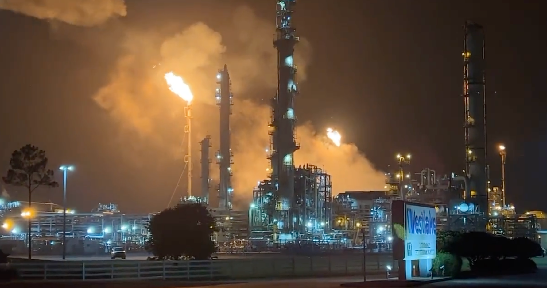 Night view of an oil refinery with multiple tall smokestacks emitting flames and smoke, illuminated by industrial lights, with a nearby sign for a business named Wal-Mart.