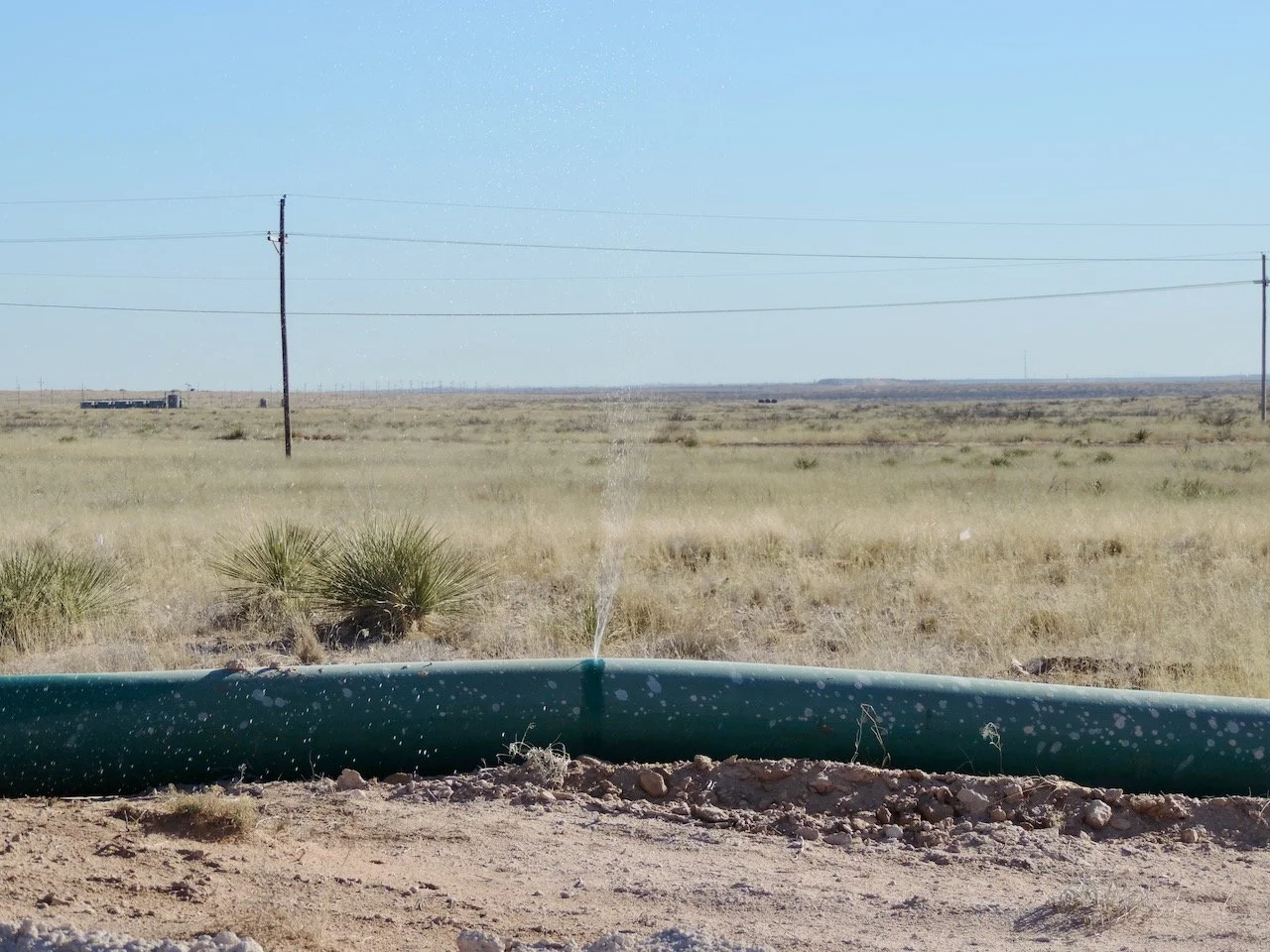 A flat, open desert landscape with sparse vegetation, power lines, and a water pipe in the foreground, under a clear blue sky.