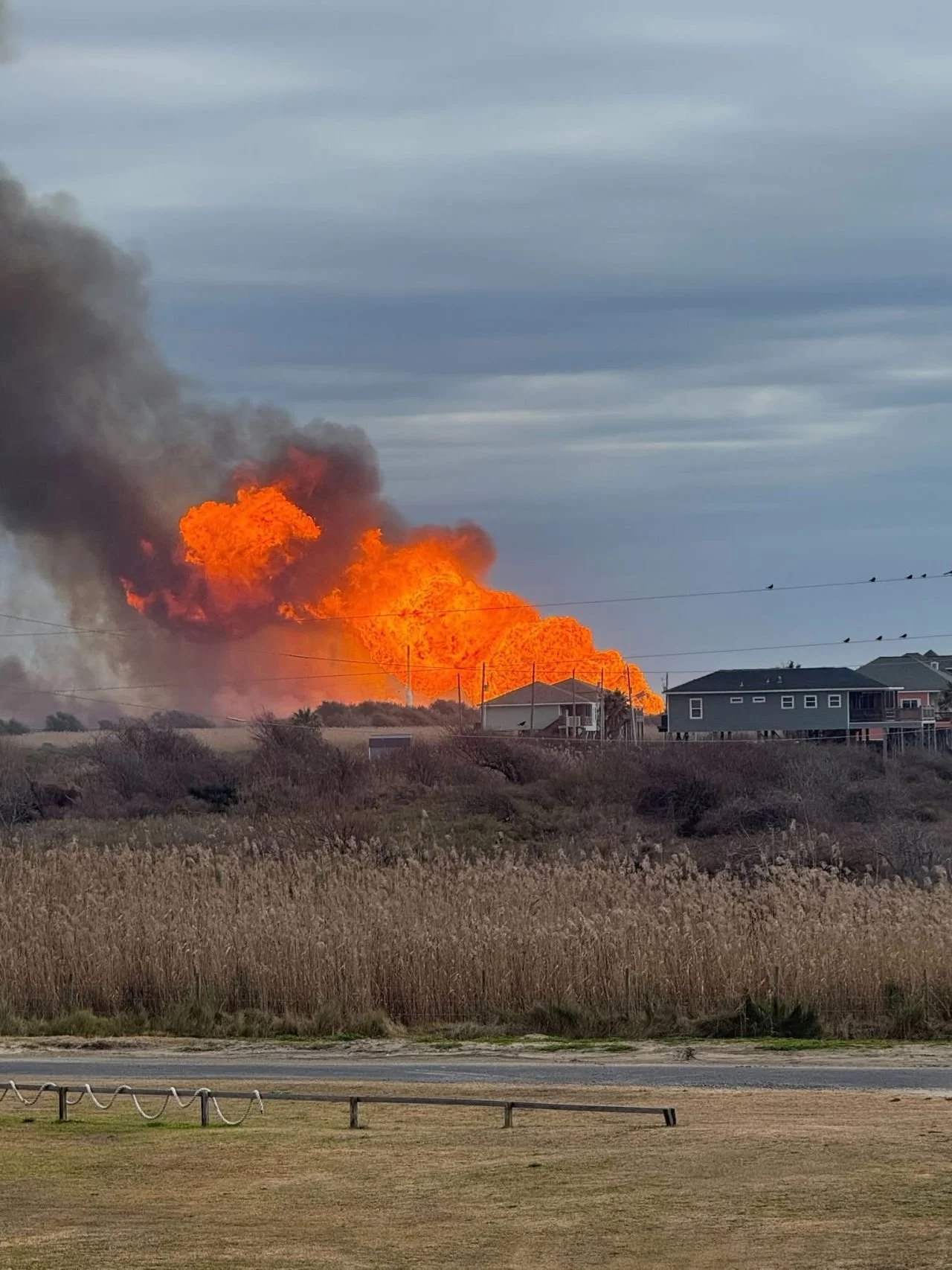 Large wildfire with bright orange flames and dark smoke rising behind residential houses in a suburban area, under a cloudy sky.