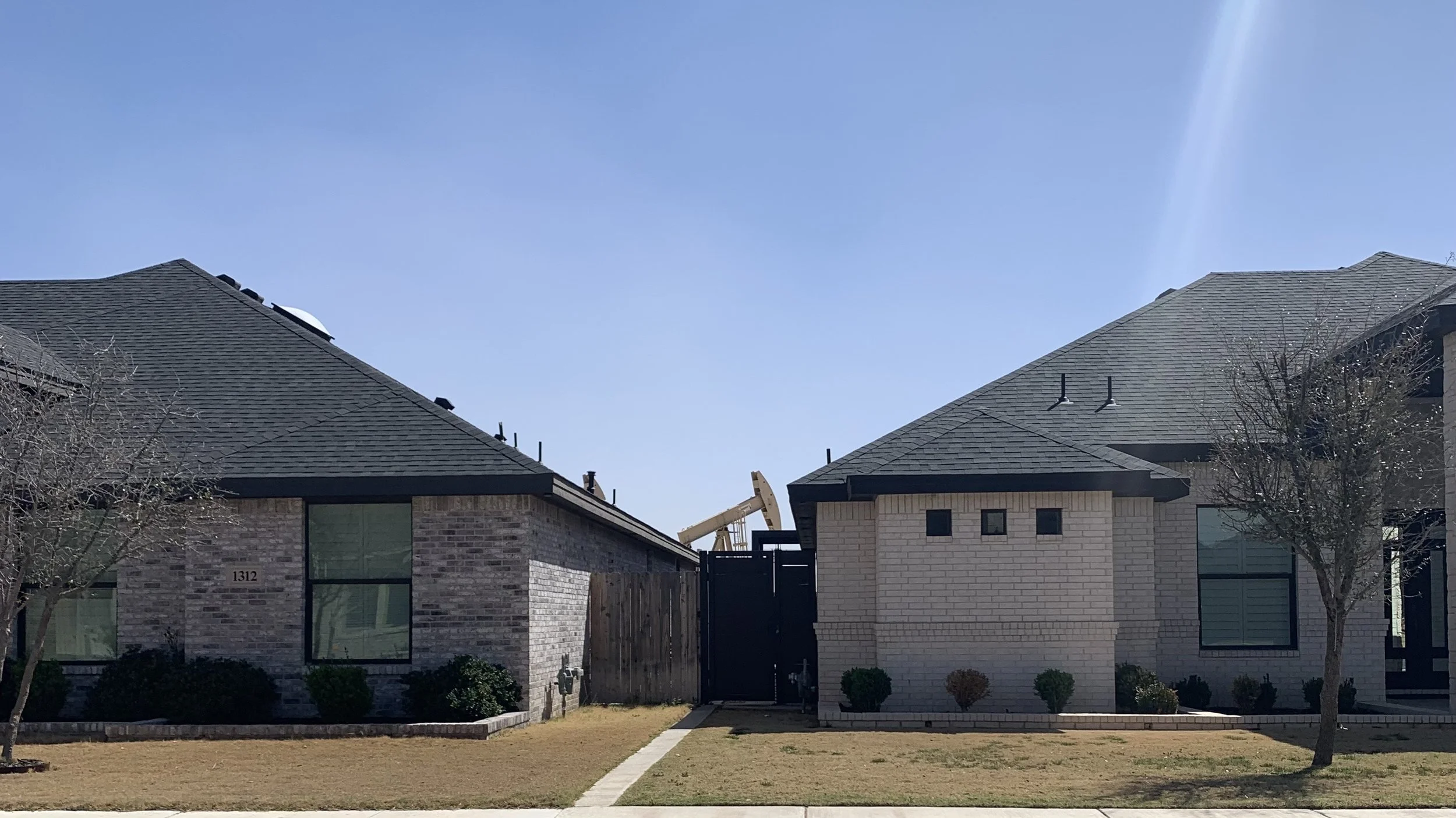 Two houses with brick exteriors and dark gray roofs, a wooden fence, a small grassy yard, trees, and a clear blue sky with sunlight.