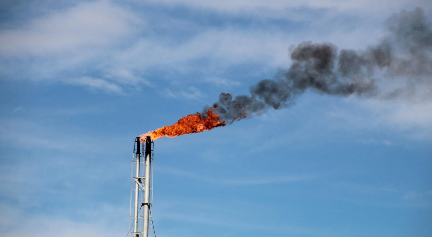 Industrial smokestack emitting fire and black smoke into a blue sky with clouds.