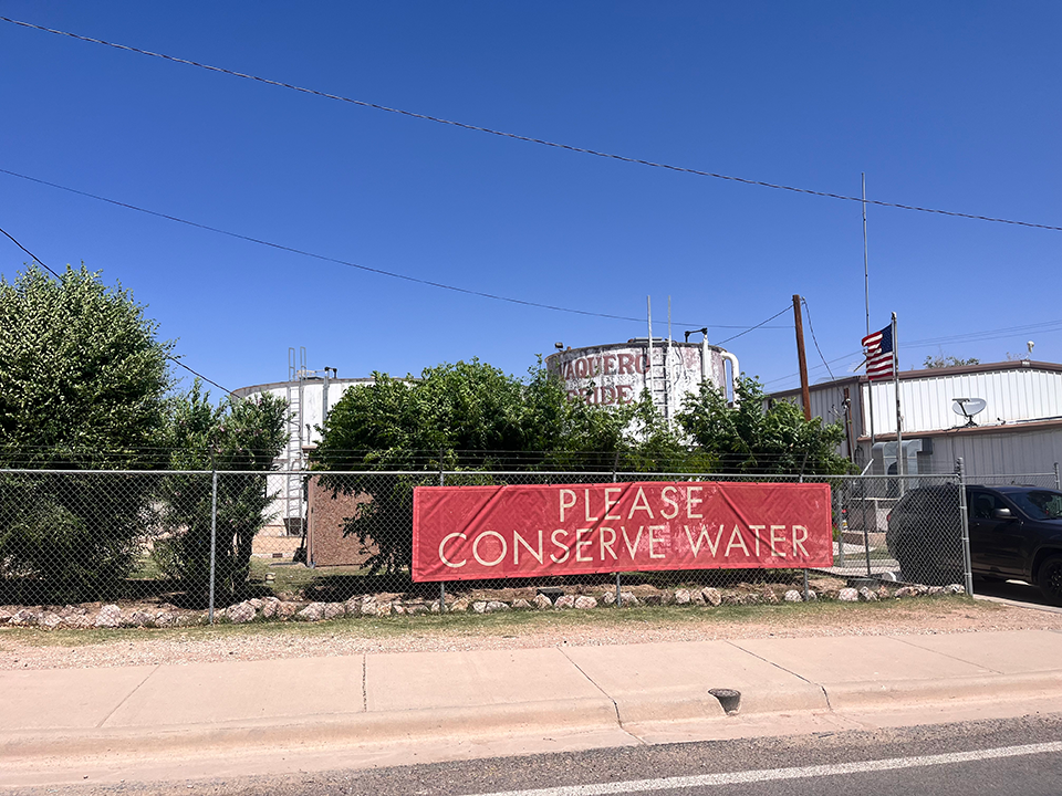 A banner on a fence says "Please Conserve Water"