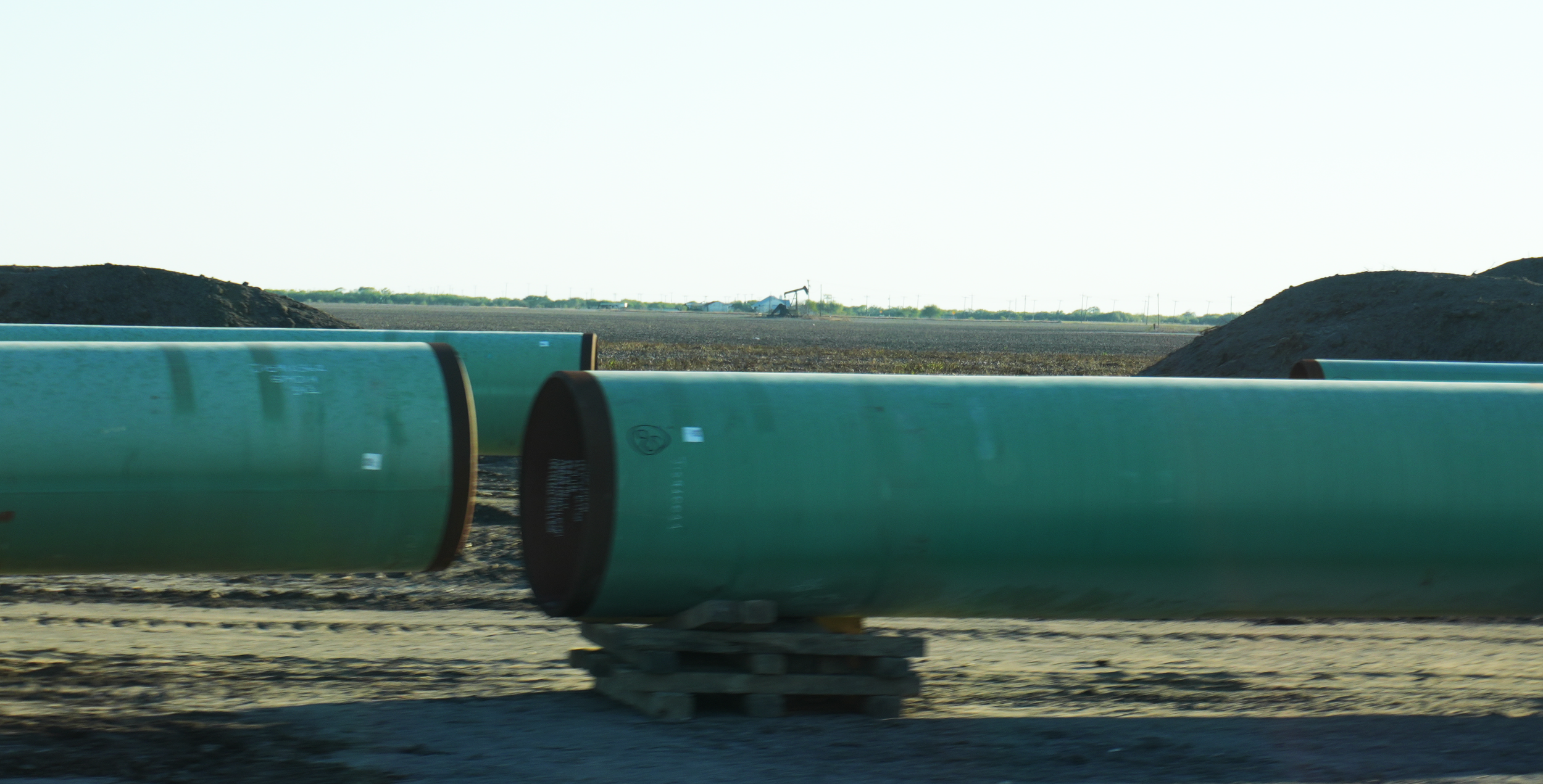 Construction pipes lying on the ground on a construction site with dirt and soil, with a view of open land and a distant oil pumpjack in the background.