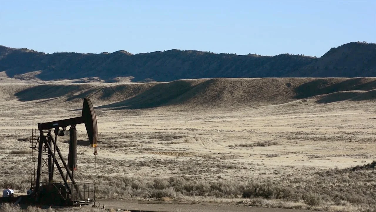 An oil pumpjack operating in a vast, open desert landscape with hills and mountains in the background under a clear sky.