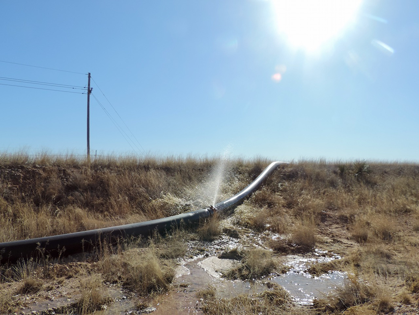 A large black pipe leaking water in a dry, grassy field under a bright blue sky with the sun shining.
