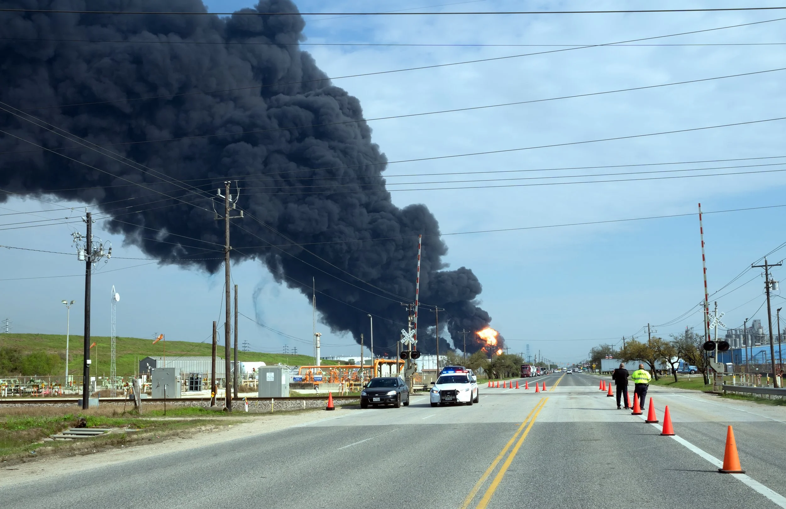 Large black smoke billowing from a fire at an industrial site, with orange flames visible, while a police car and emergency personnel are present on a nearby road lined with orange cones and railroad crossing signals.