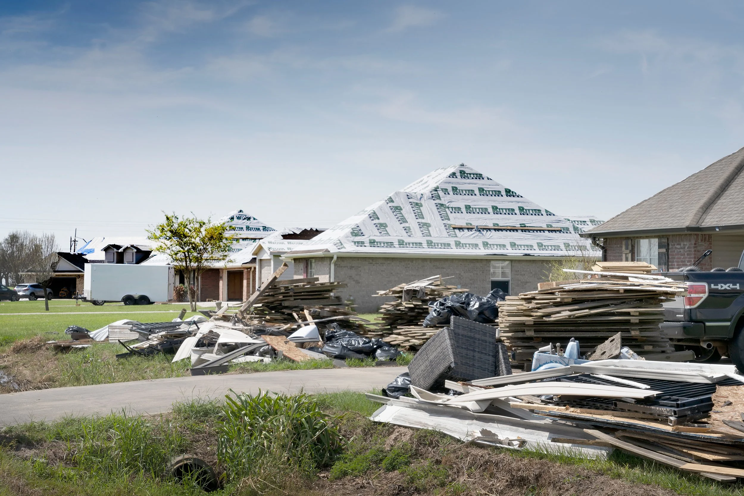A house under construction with shingles partially installed on the roof, surrounded by construction materials and debris, with a clear blue sky in the background.