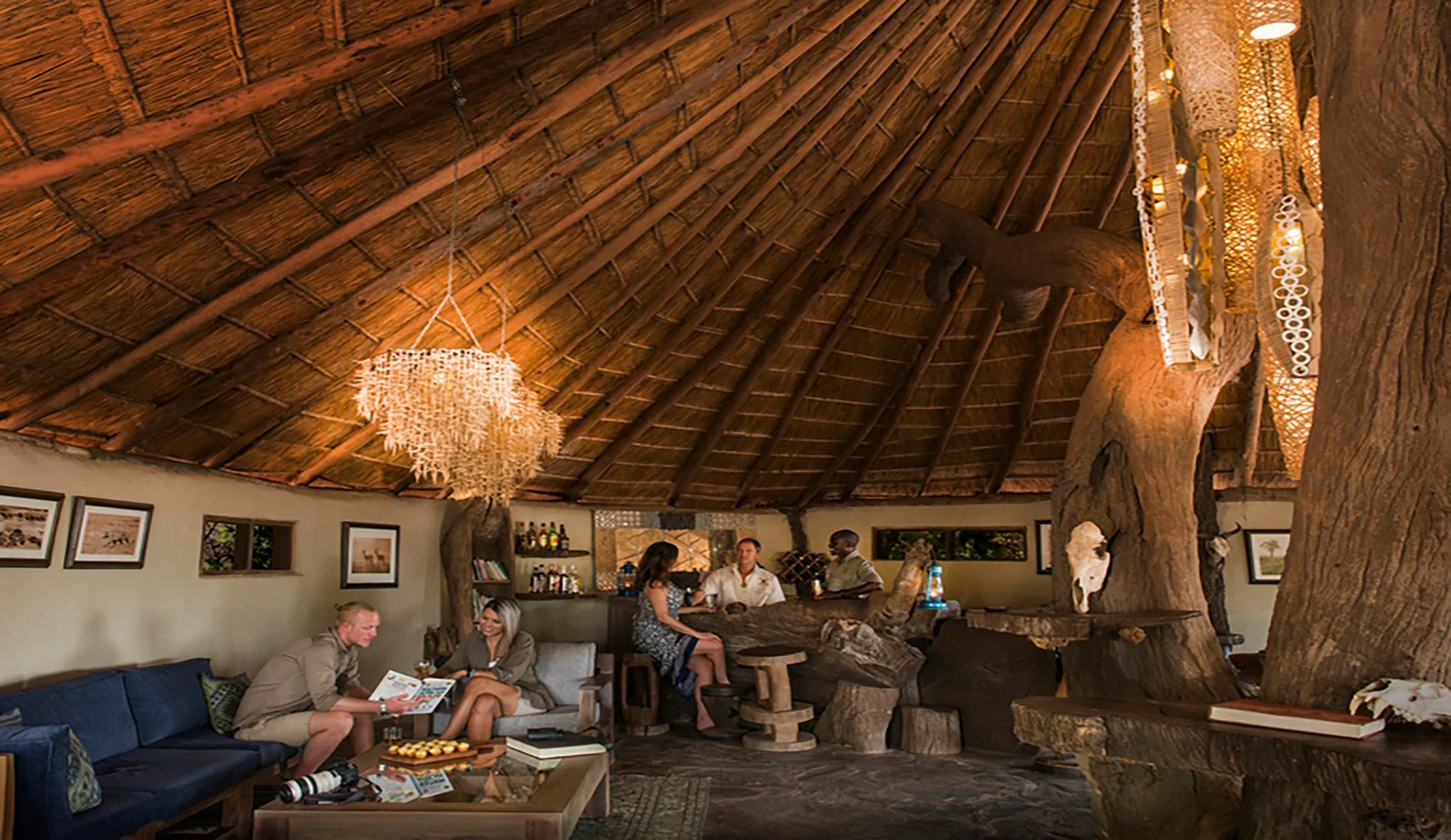 Interior of a tropical-themed restaurant or bar with a thatched roof, wooden decor, and modern chandeliers. Several people are seated and socializing near a bar area. Kaingo camp. Safari in South Luangwa national park with Wild Selection Tours.
