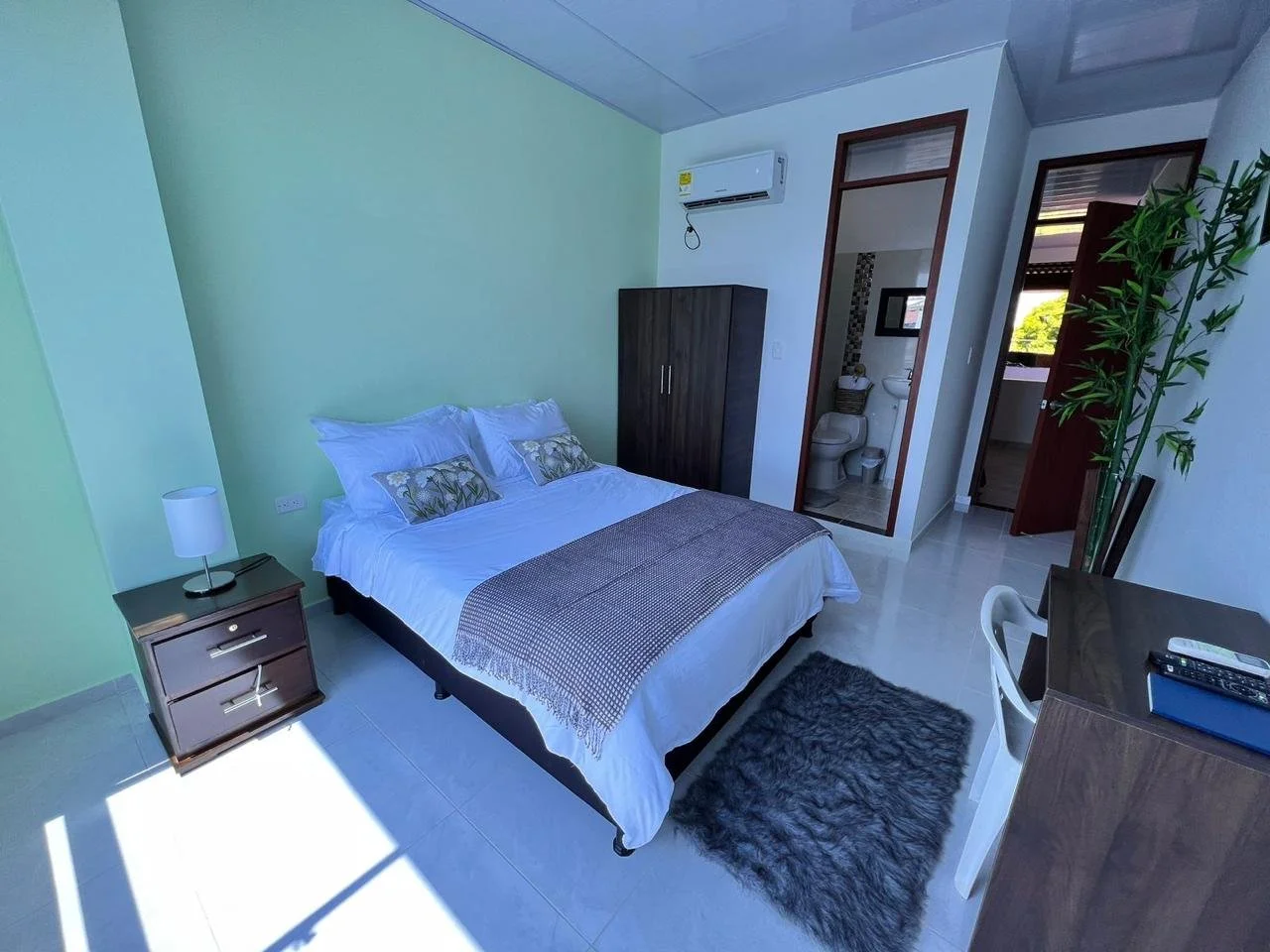 Hotel room with a bed, nightstand, lamp, wardrobe, desk with a chair, and a small furry rug on the floor. The room has a green accent wall and an air conditioning unit. There is a bathroom visible through an open door.