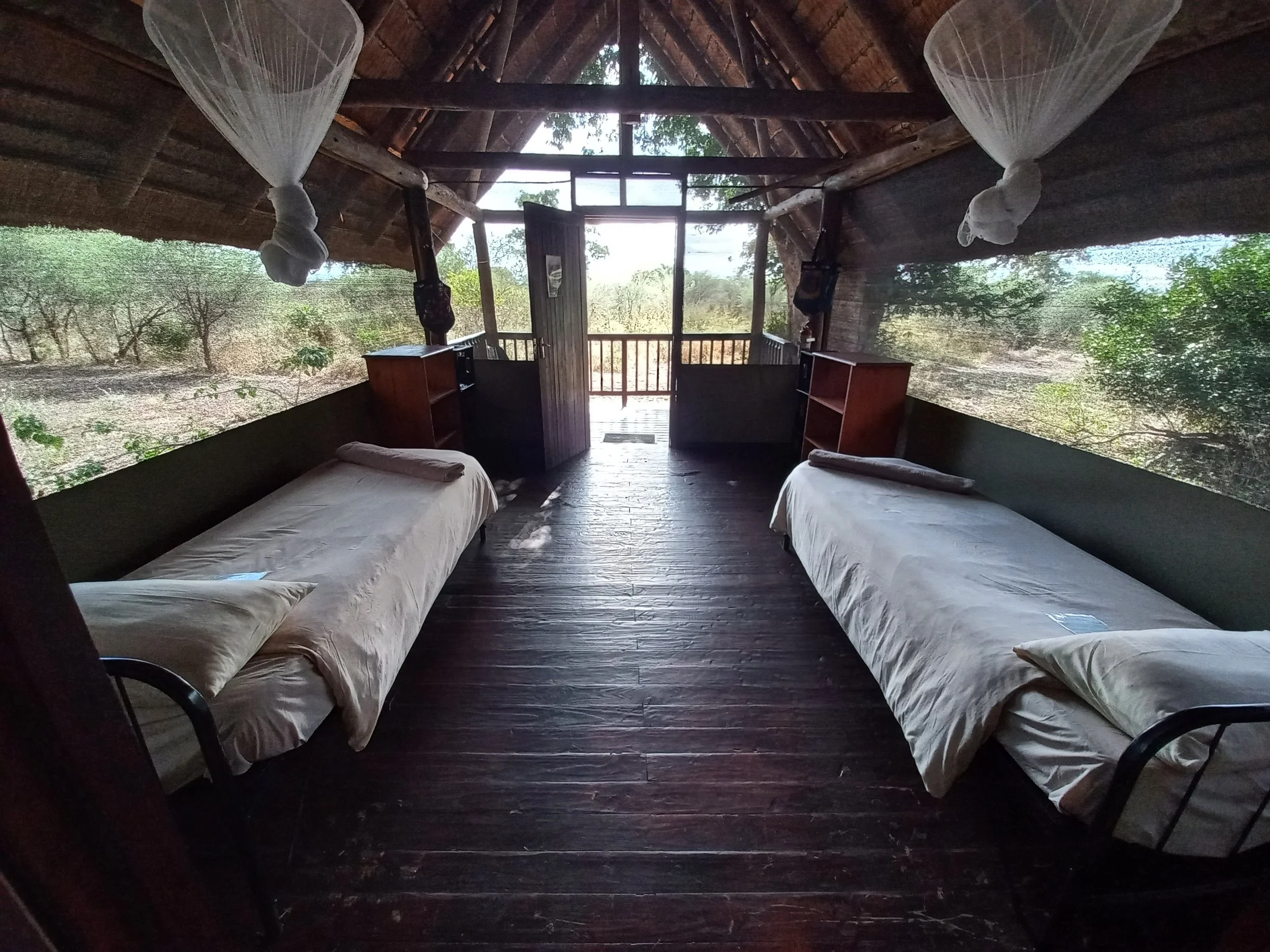Interior of a rustic cabin with two single beds covered with white linens, positioned on either side. The cabin has a wooden floor and bamboo walls, with large open windows showing a natural outdoor landscape. There's a small balcony with a wooden railing at the far end, and two hanging mosquito nets are above each bed.