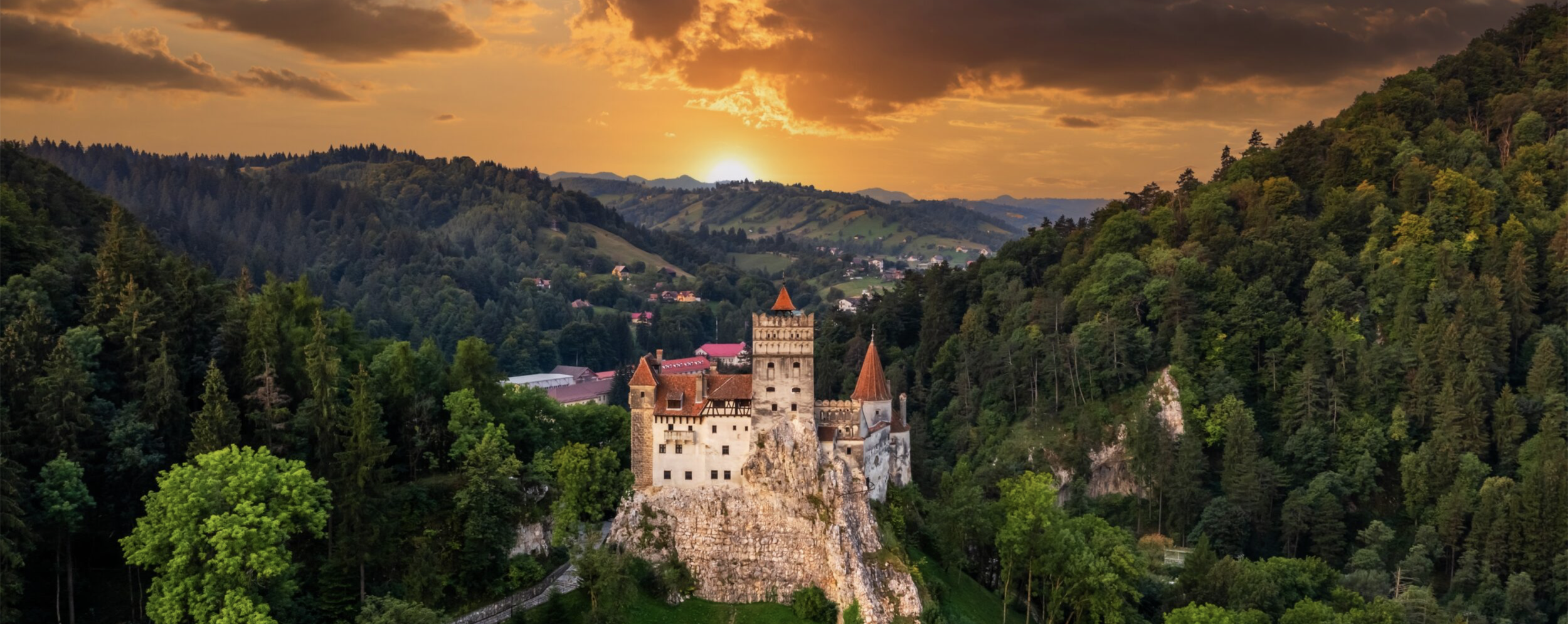 A medieval castle on a rocky hill surrounded by dense green forest with a sunset sky in the background.