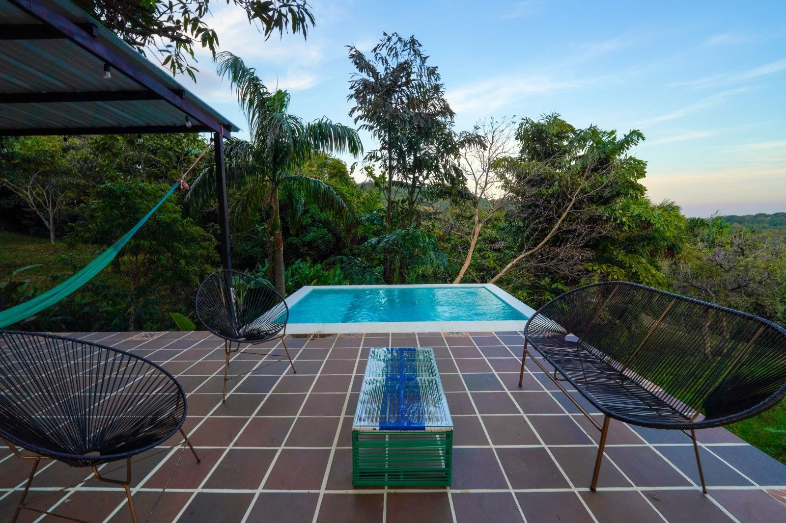 Outdoor patio with black wire chairs, a small table, a hammock, and a small pool surrounded by greenery and trees under a blue sky.