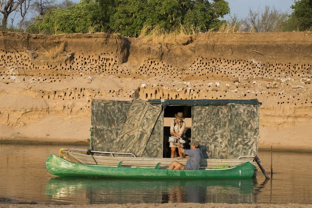 A person in a kayak on a calm river is taking a photograph of another person inside a floating structure with camouflage fabric walls, situated on the river, with a dirt embankment and trees in the background.Kaingo camp. Safari in South Luangwa.