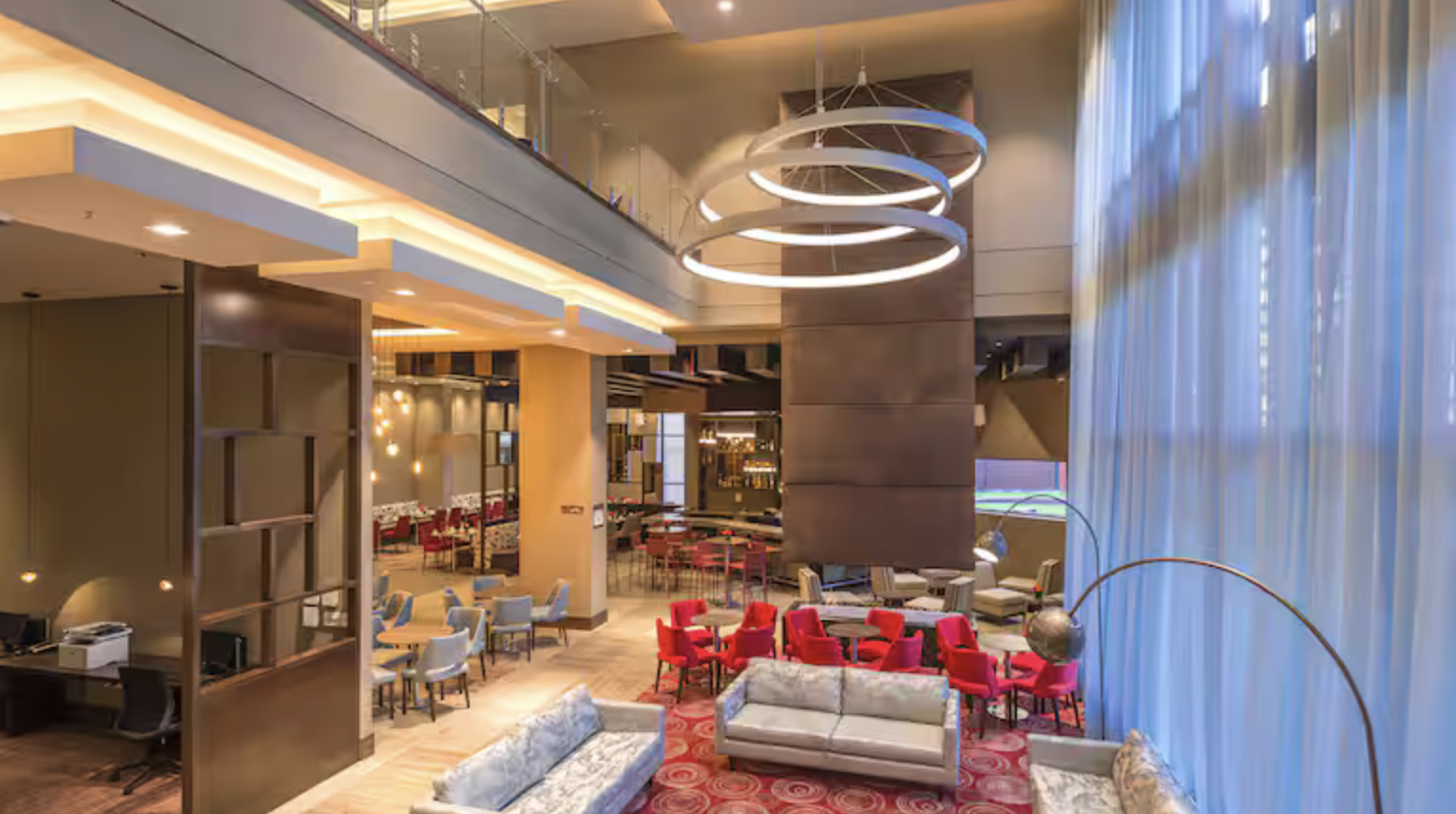 Interior of a modern hotel lobby with a high ceiling, circular chandeliers, colorful chairs, sofas, and large floor-to-ceiling curtains near a wall of windows.