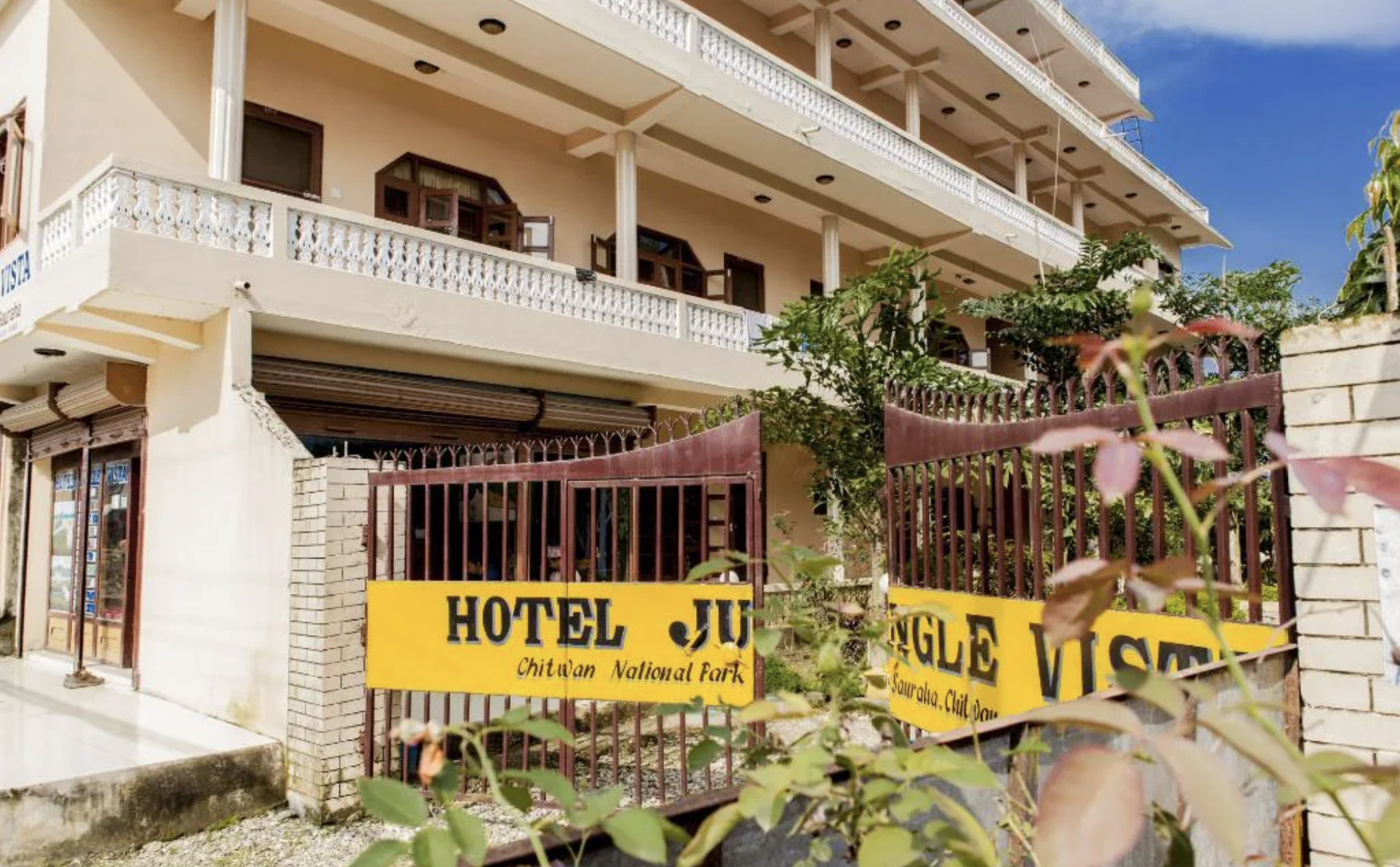 Multi-story hotel building with white balconies and a brown gate. Yellow signage on the gate reads 'HOTEL JUNGLE, Chitwan National Park, Sauraha, Chitwan' in black text. Wildlife expedition and phot safari with Wild Selection Tours.