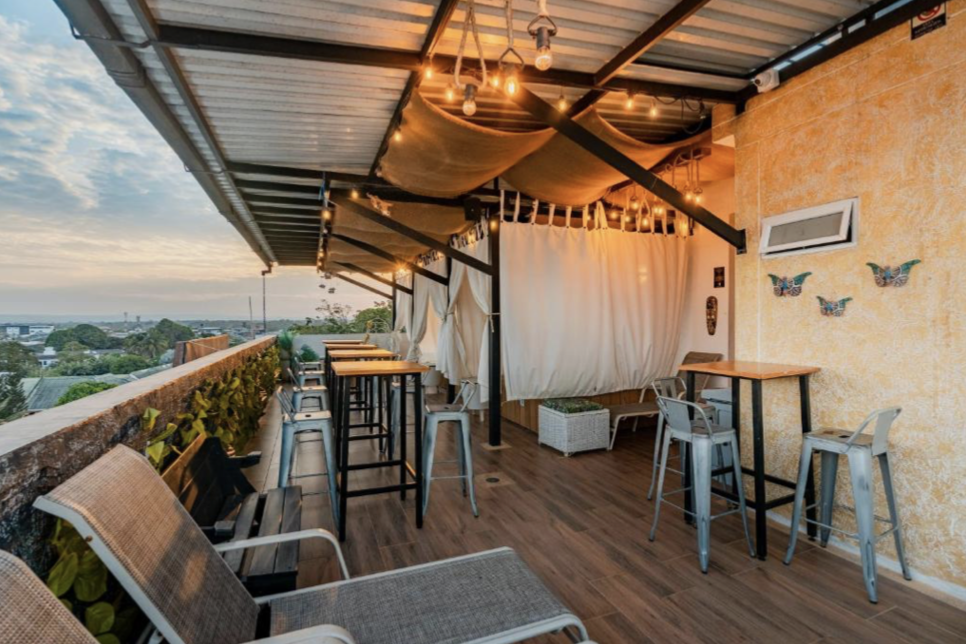 A rooftop terrace with outdoor seating, high-top tables, bar stools, decorative butterfly wall art, string lights, and a view of the cityscape, during sunset.