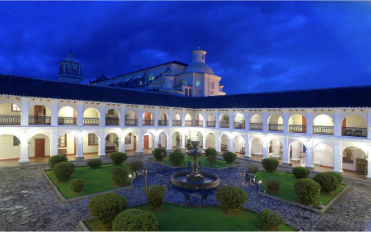 A courtyard with a central fountain surrounded by trimmed bushes, stone paths, and a two-story colonial-style building with arches and balconies, illuminated at dusk.