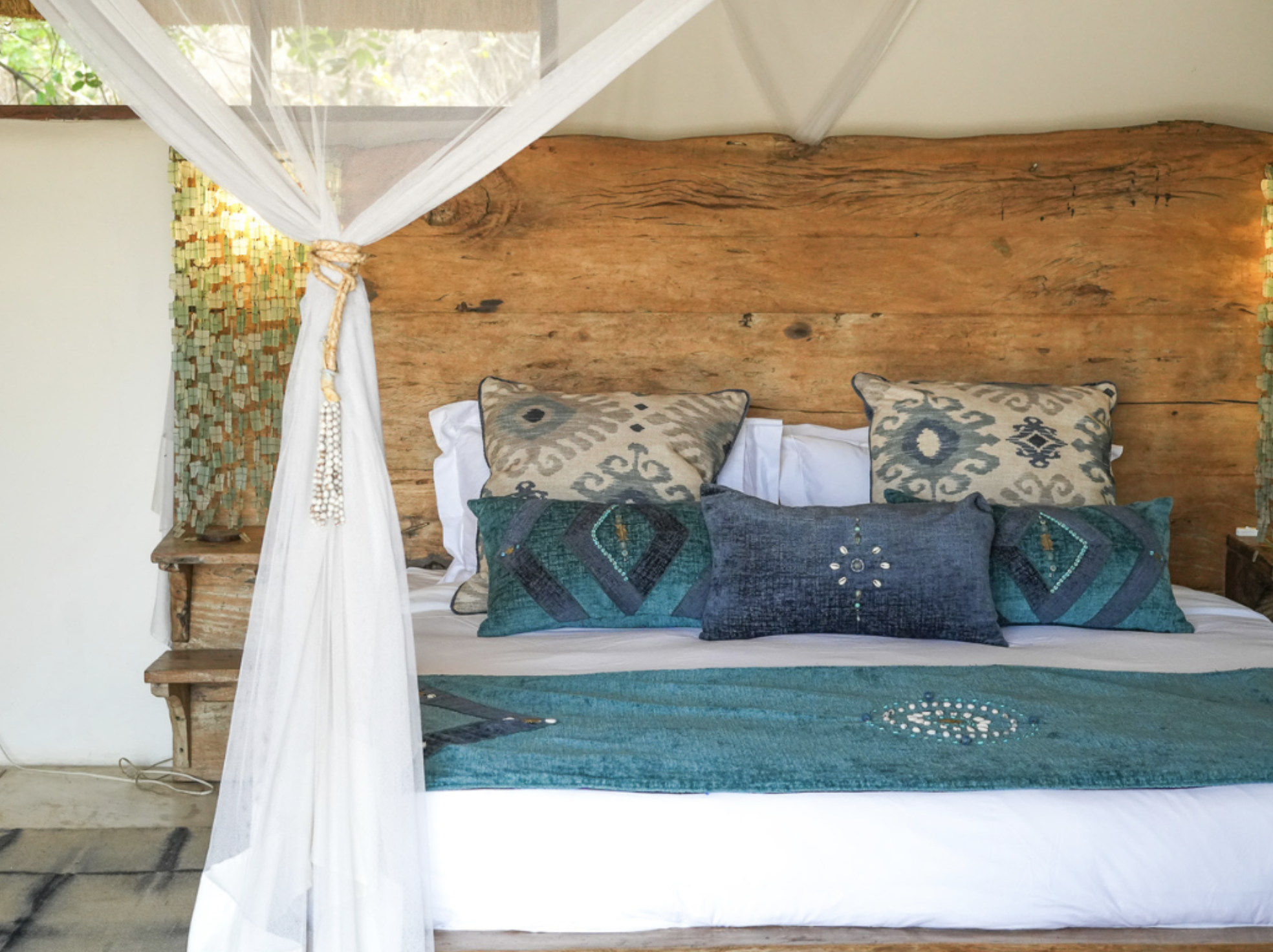 A bed with a rustic wooden headboard, white bedding, and decorative pillows in beige, gray, and teal. A white canopy drapes over the bed, tied to the posts, and a wooden side table is visible on the left.Kaingo camp. Safari in South Luangwa, Zambia.