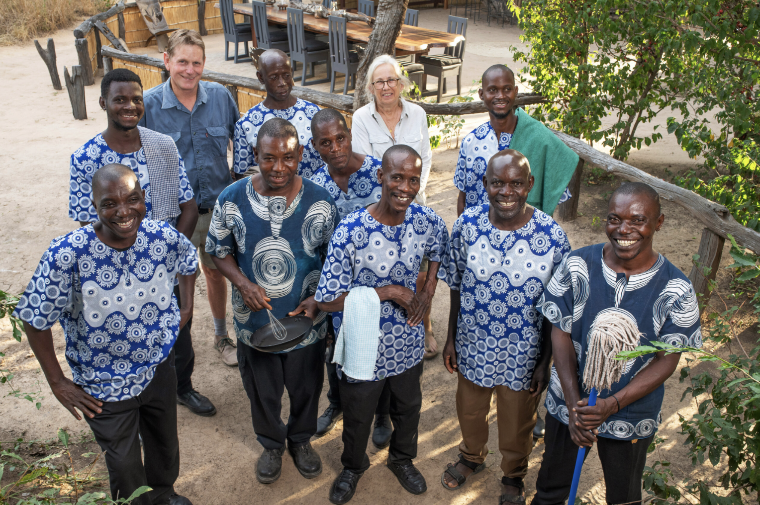 A group of people, including men and women, gathered outdoors on a dirt area, smiling at the camera. They are wearing traditional patterned clothing, with some holding tools. In the background, there are trees, a wooden fence, and a dining area with chairs and tables.