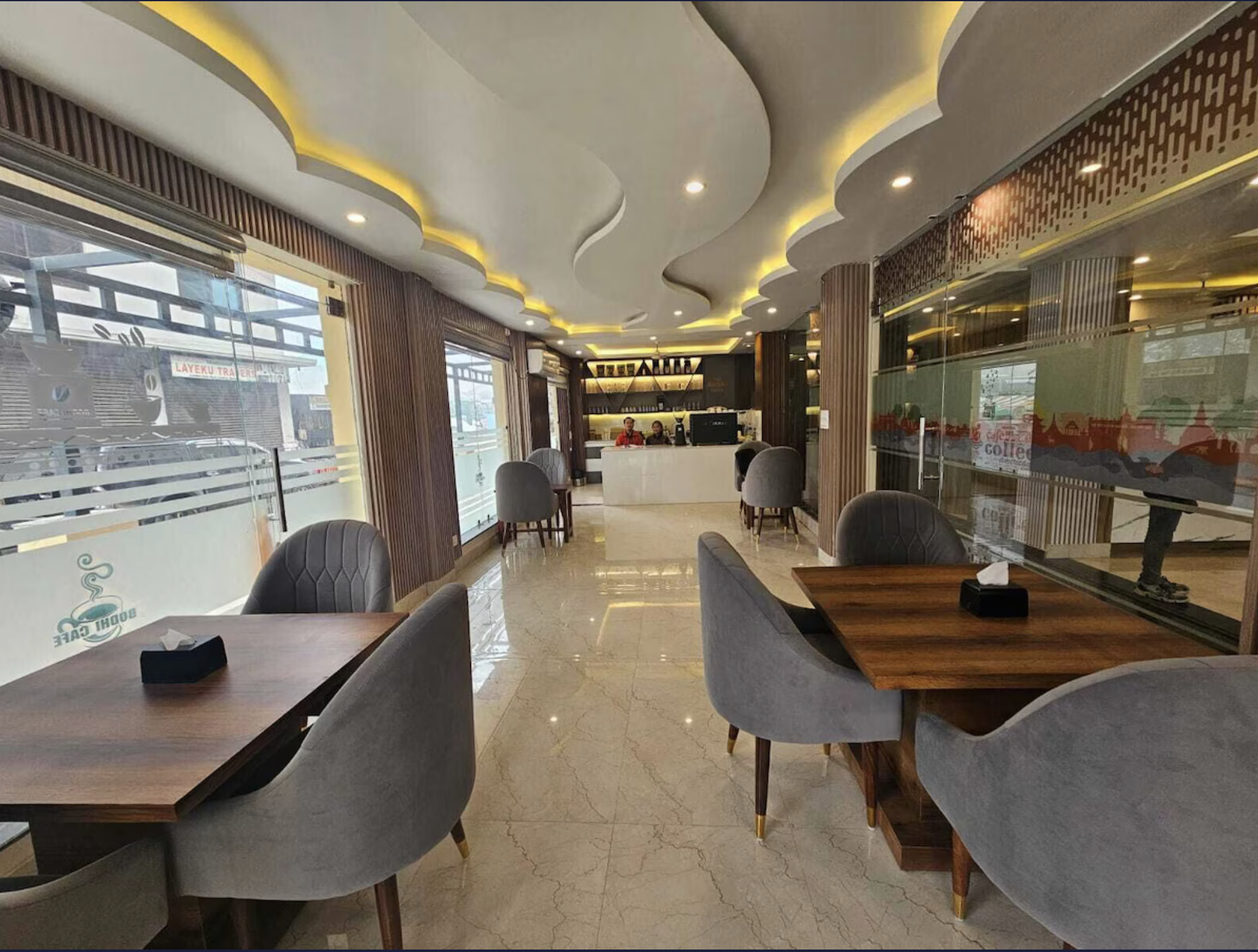 Inside a modern cafe with marble flooring, wood accents, and large glass windows. There are several tables with gray upholstered chairs and a white counter at the back with people working. Wildlife expedition and phot safari with Wild Selection Tours