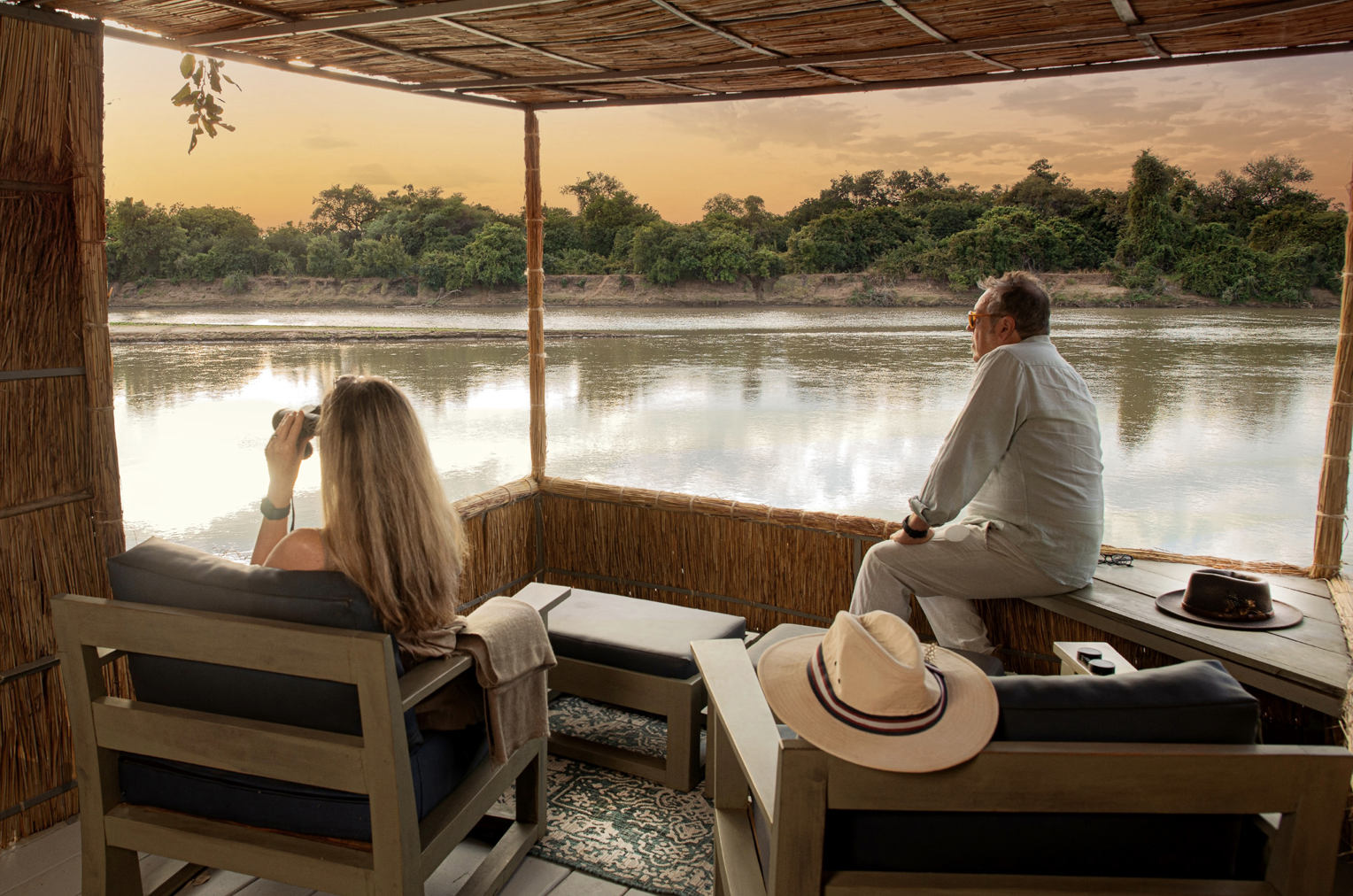 Two people sitting on a boat or floating hut looking out over a river at sunset, with lush green trees on the opposite bank. Kaingo camp. Safari in South Luangwa national park with Wild Selection Tours.