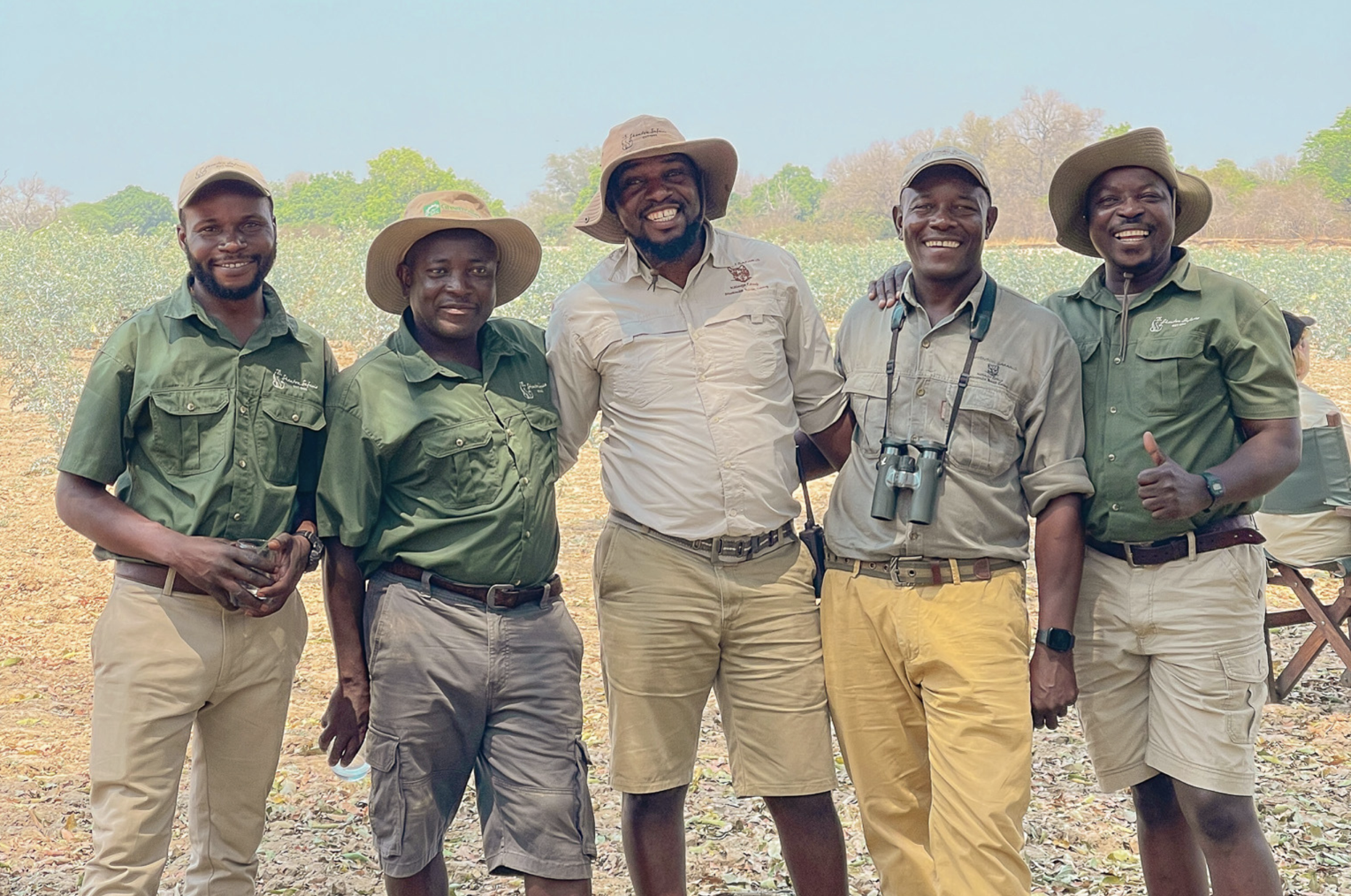 Group of five Zambian gui standing outdoors in a field, smiling, dressed in safari attire, some wearing hats, one with binoculars around his neck, and one holding a camera. Kaingo camp. Safari in South Luangwa national park with Wild Selection Tours.
