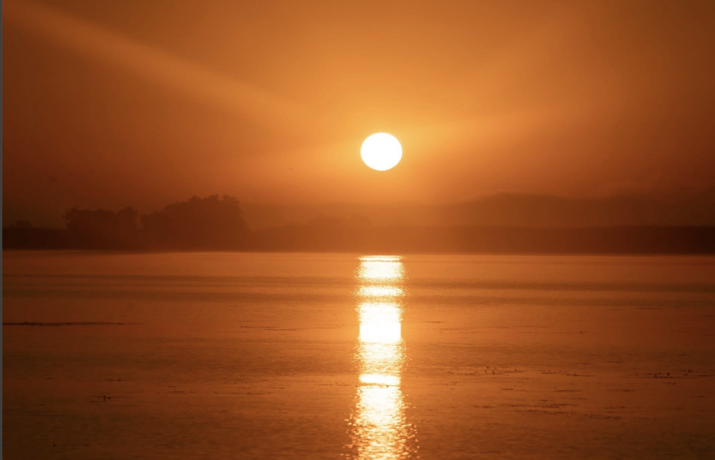 Sunset over a body of water with orange sky and silhouetted land in the distance.