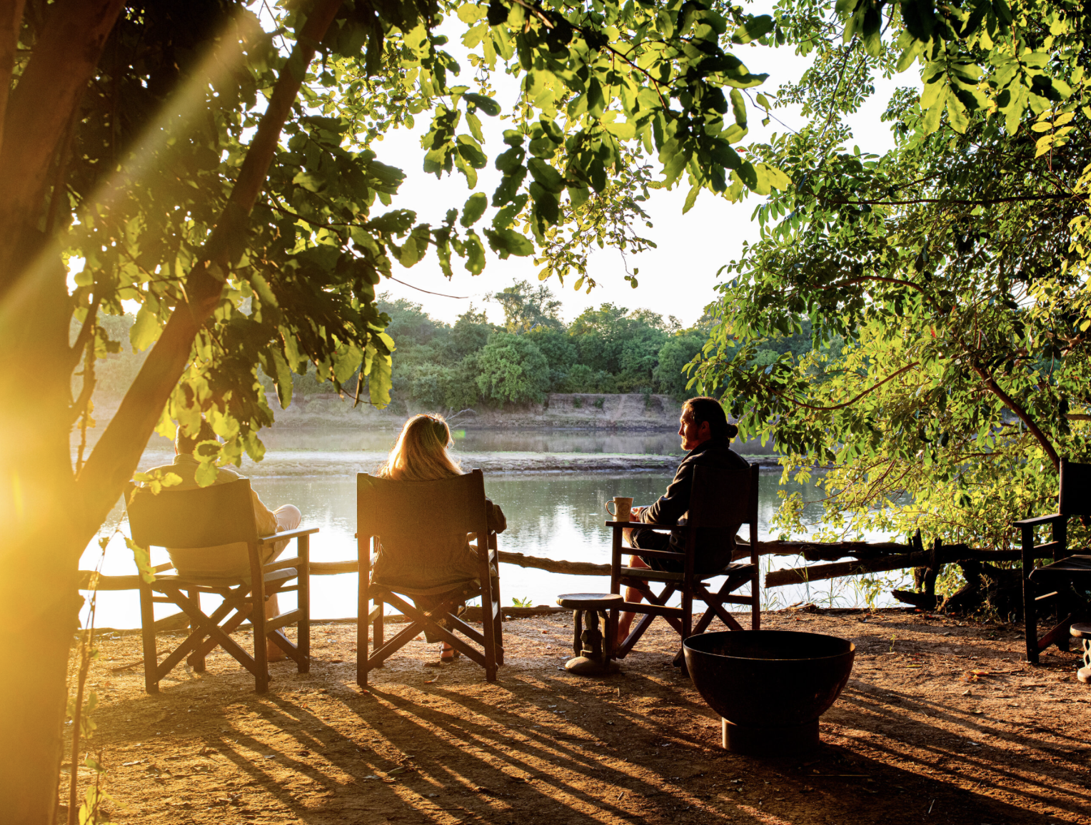 Three people sitting on outdoor chairs by a river, surrounded by trees with sunlight filtering through their leaves. Kaingo camp. Safari in South Luangwa national park with Wild Selection Tours.