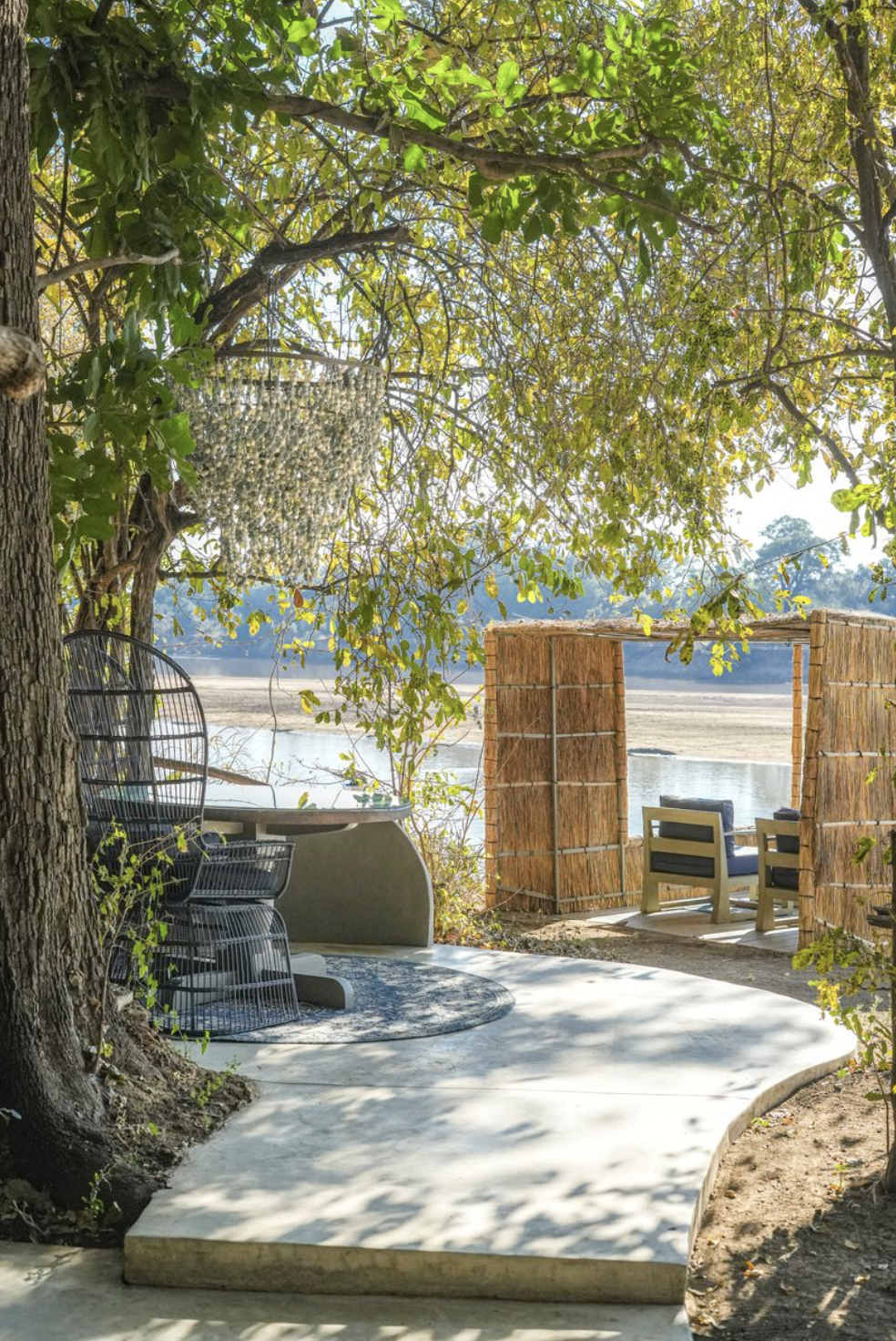 A lakeside outdoor seating area with a shaded patio under trees. Includes a round table with black wire chairs, and a bamboo privacy screen with two chairs facing the lake beyond. The scene is sunny with reflections on the water. Kaingo camp.