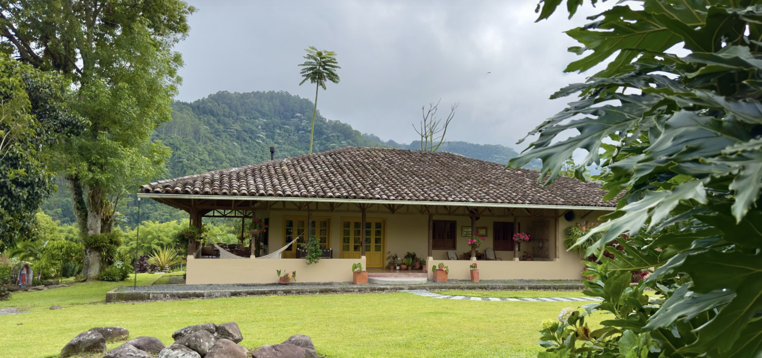 Single-story house with a tiled roof, yellow doors, and surrounded by lush greenery and potted plants in a rural setting with mountains and cloudy sky in the background.