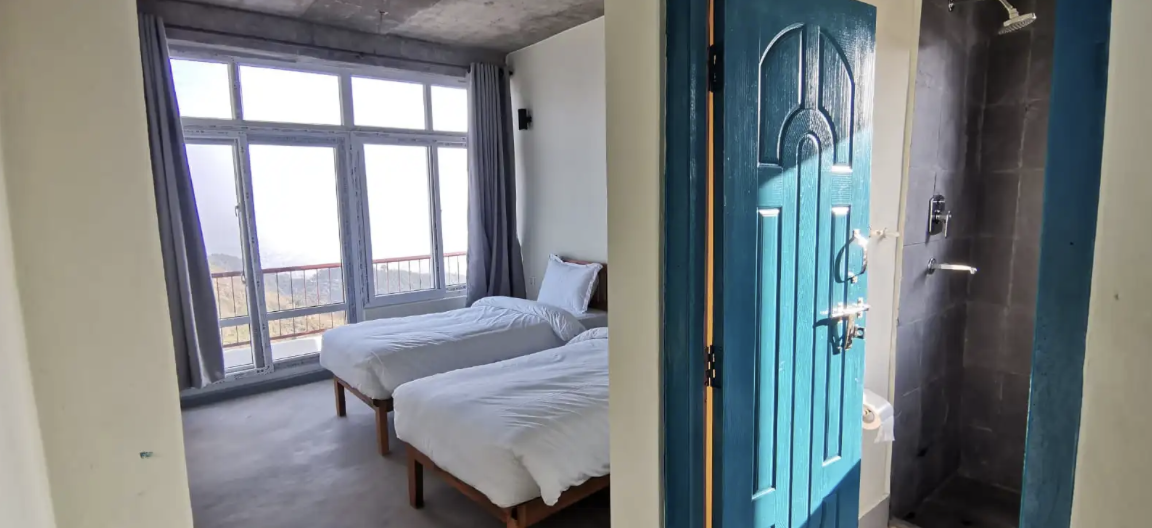 Hotel room with two beds positioned near large windows with curtains overlooking an outdoor balcony, adjacent to an open shower area with blue door. Wildlife expedition and phot safari with Wild Selection Tours.