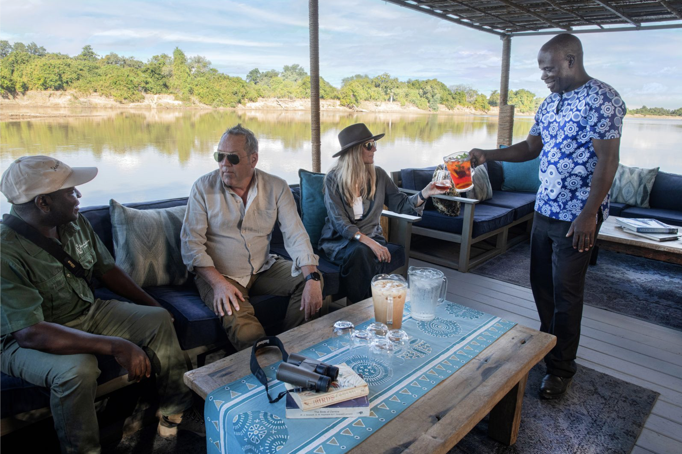 Group of four people on a boat deck overlooking a river, enjoying drinks. One man is pouring a red drink into a woman's glass, while others appear engaged in conversation. Kaingo camp. Safari in South Luangwa national park with Wild Selection Tours.