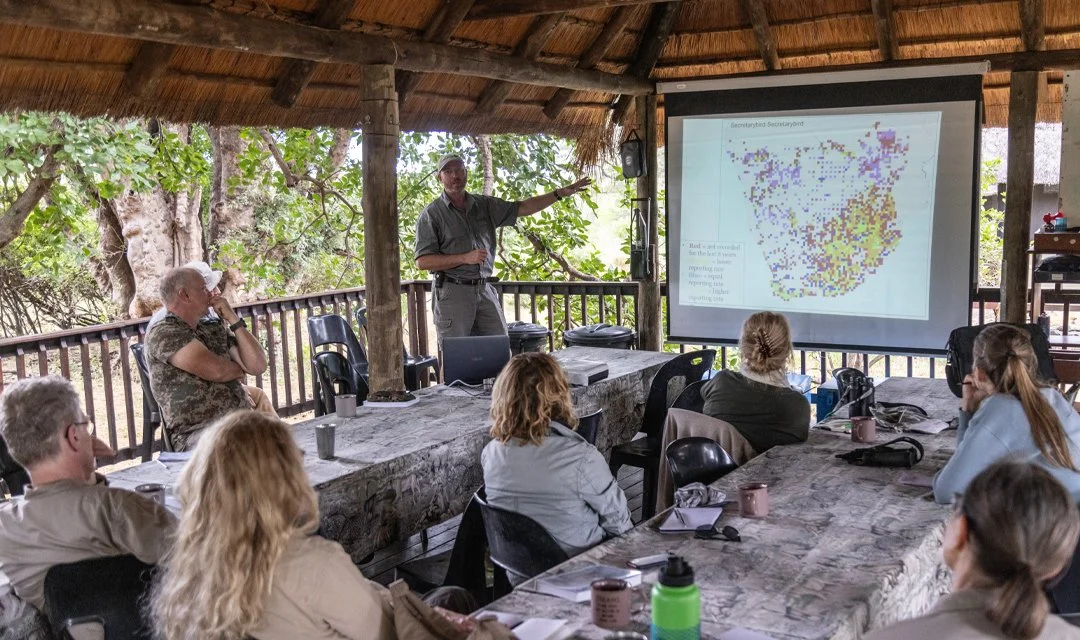 A group of people attending a presentation in a covered outdoor setting, with trees in the background, listening to a presenter pointing at a projection screen displaying a colorful map or data visualization.