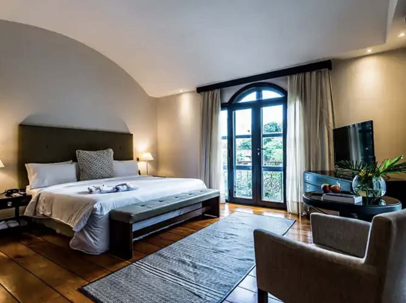 Hotel room with large bed, nightstands, a bench at the foot of the bed, a window with curtains, a flat-screen TV on a cabinet, and a small table with a vase of flowers and a bowl of fruit.