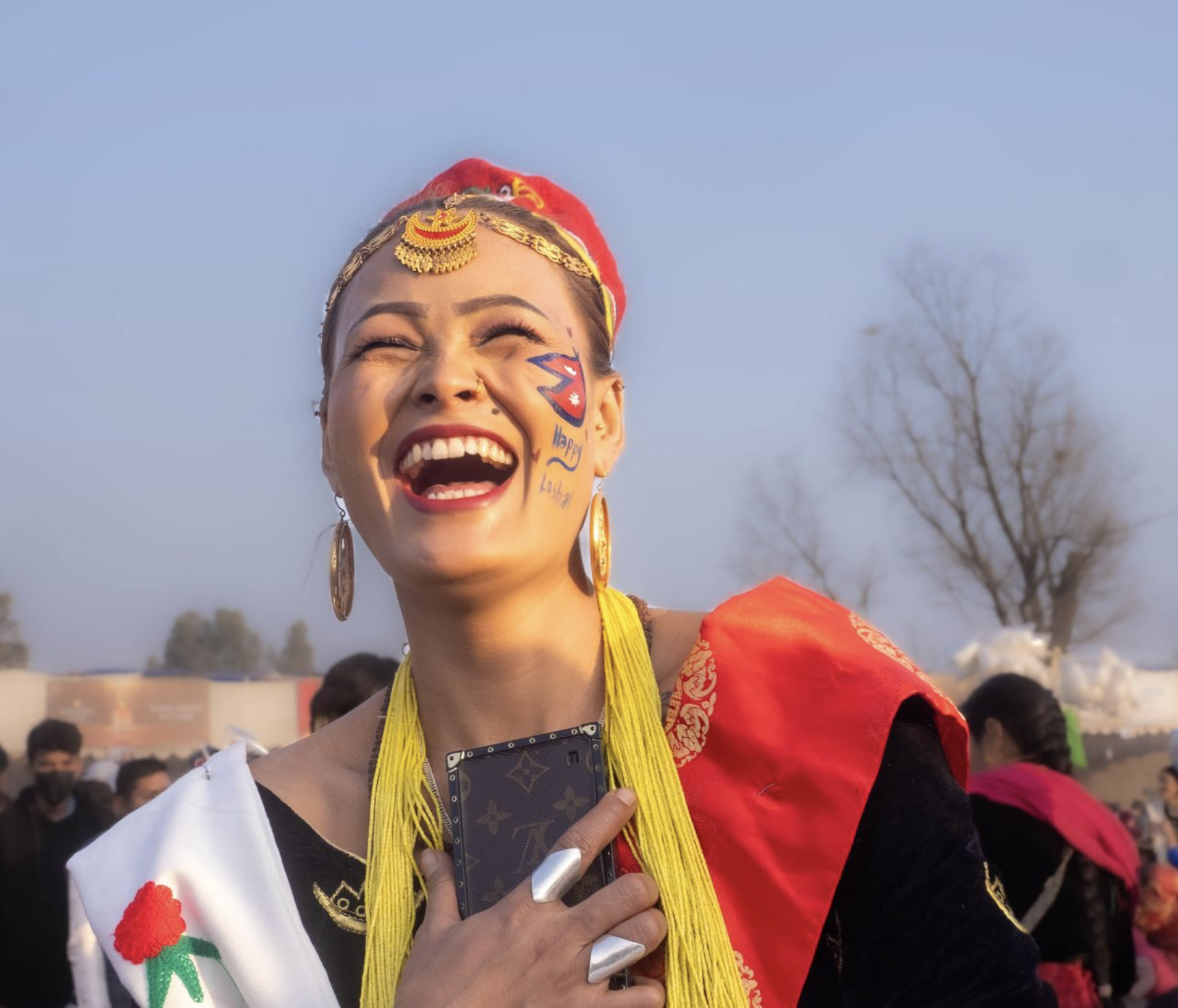 A woman dressed in traditional Nepali attire, smiling and laughing, wearing gold jewelry, a red and white scarf, and holding a smartphone. She has face paint of the Nepal flag and the words 'Happy Nepal' painted on her cheek, with a crowd and trees in the background.