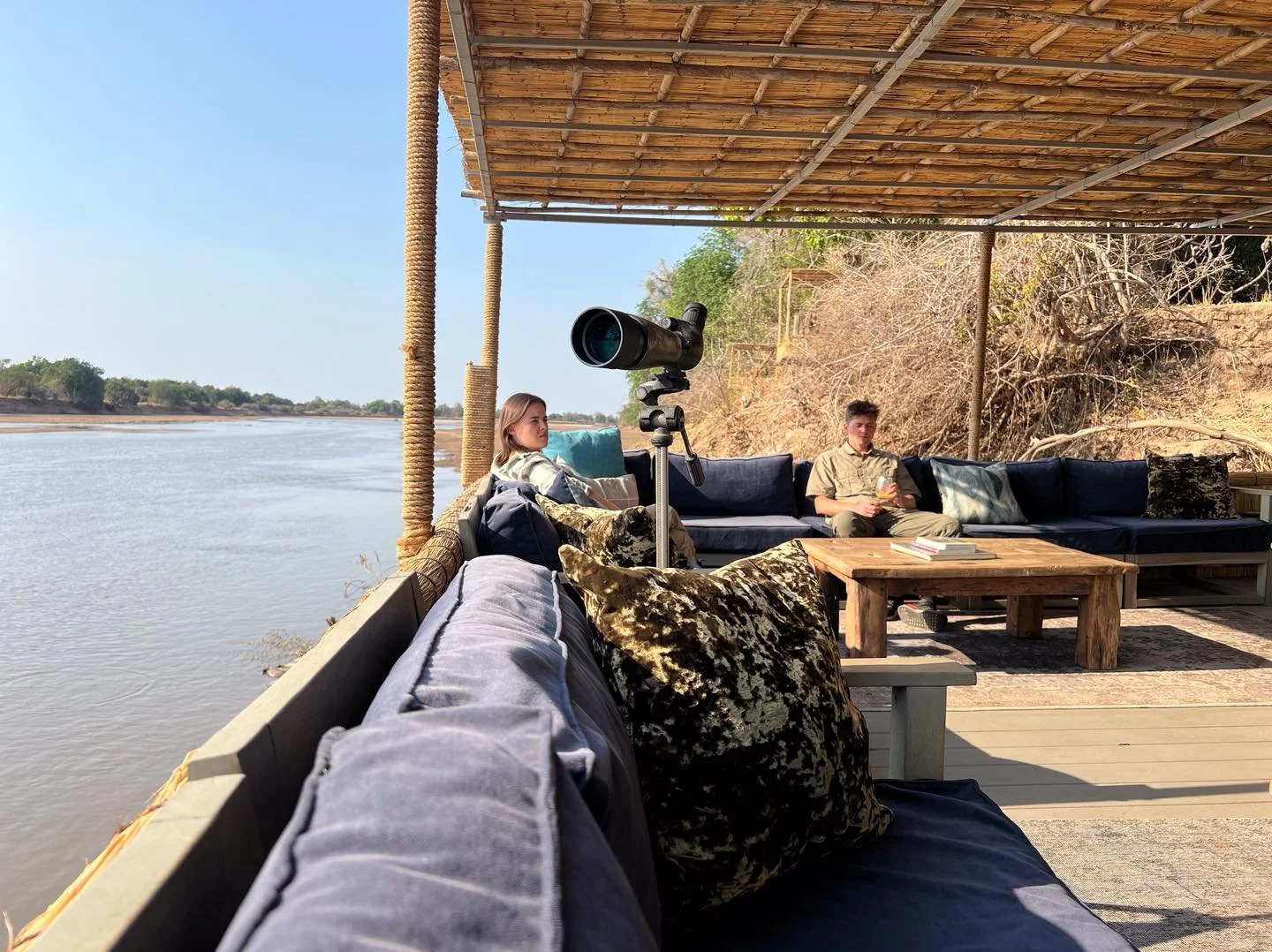 Two people sitting on an outdoor sofa under a shaded canopy by a river, with a telescope set up nearby, surrounded by cushions and rustic furniture. Kaingo camp. Safari in South Luangwa national park with Wild Selection Tours.